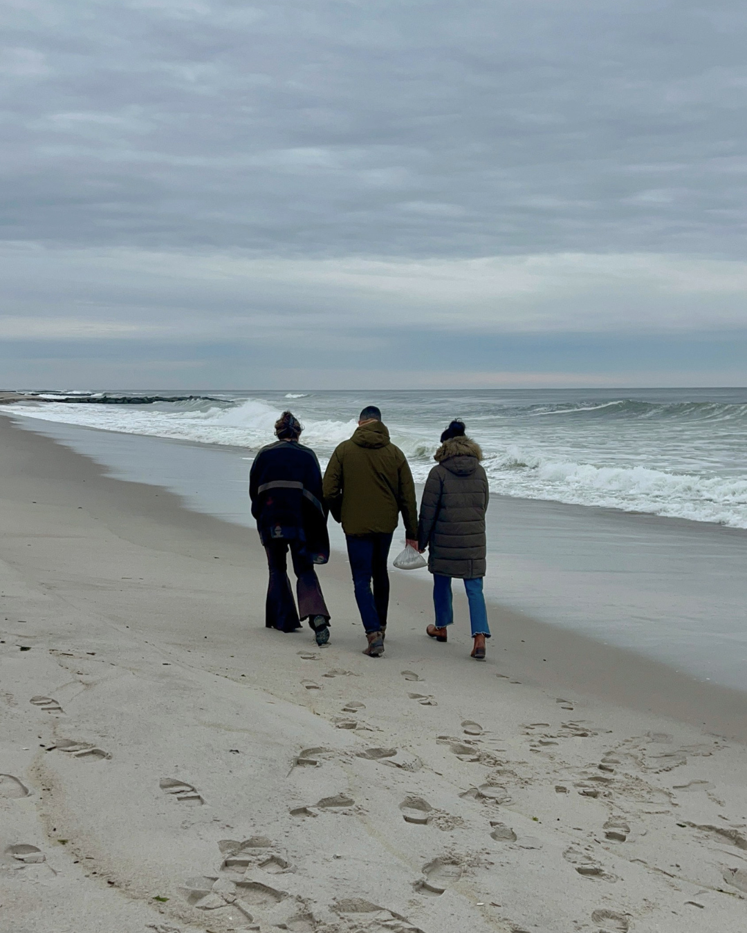 Three people walking along a sandy beach on a cloudy day, with footprints in the sand and waves in the ocean.
