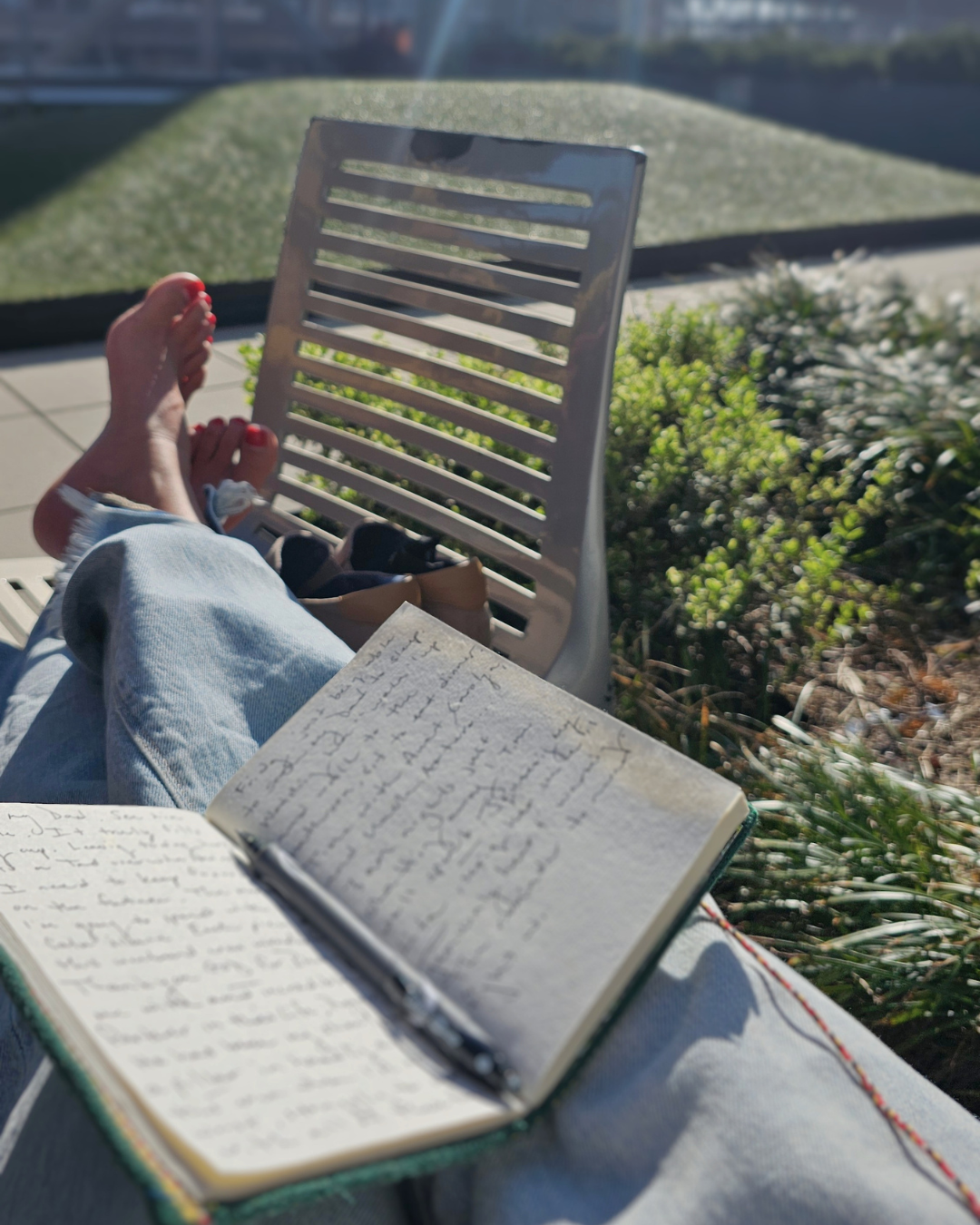 Person relaxing outdoors with feet up, writing in a notebook, with a pastel-colored backpack nearby, on a sunny day.