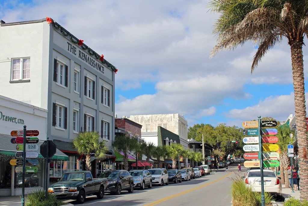 Street view in downtown with parked cars, a palm tree, commercial buildings, and a signpost with various business signs.