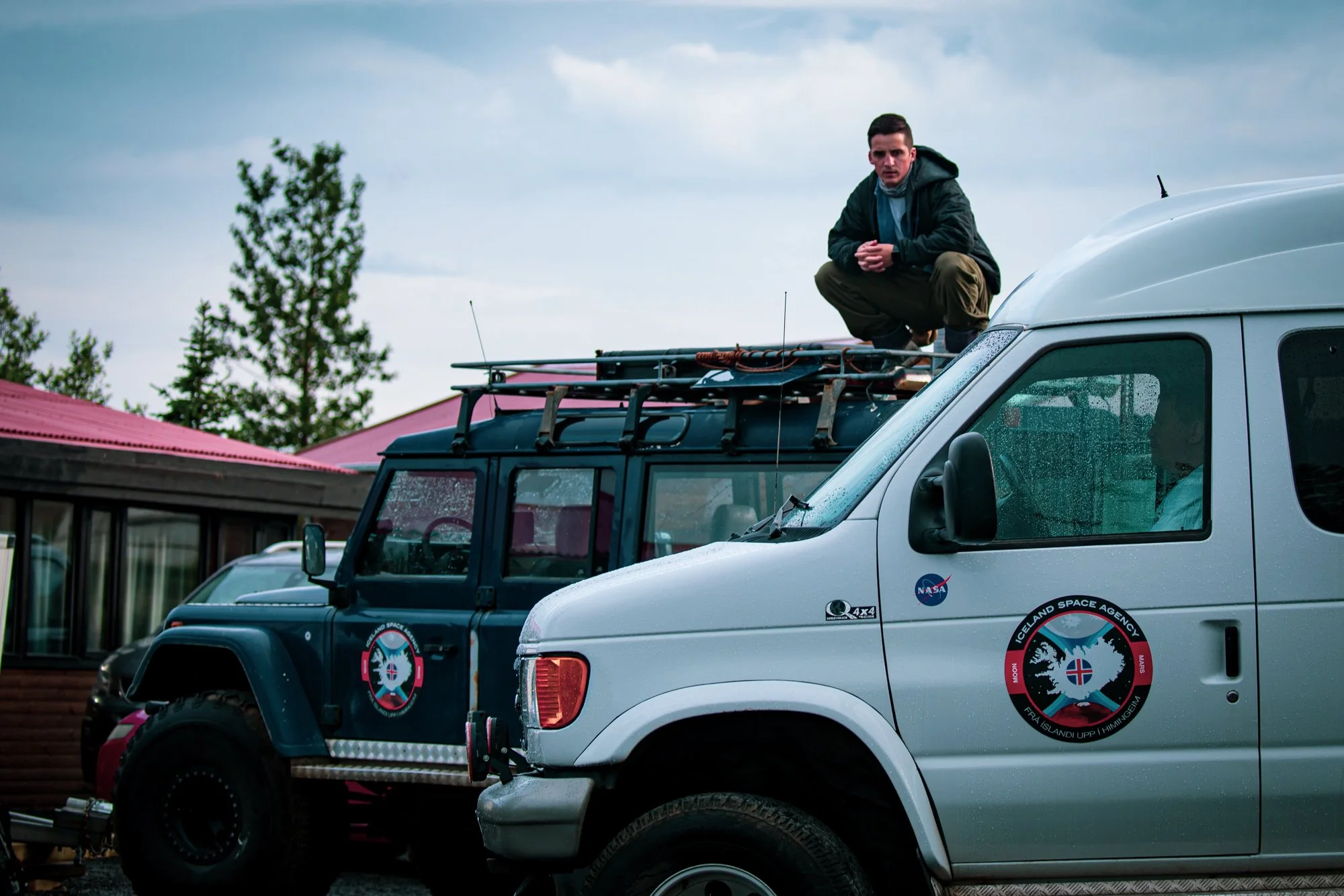 A young man crouching on top of a vehicle with Icelandic Space Agency and NASA logos, parked in front of a building with trees in the background