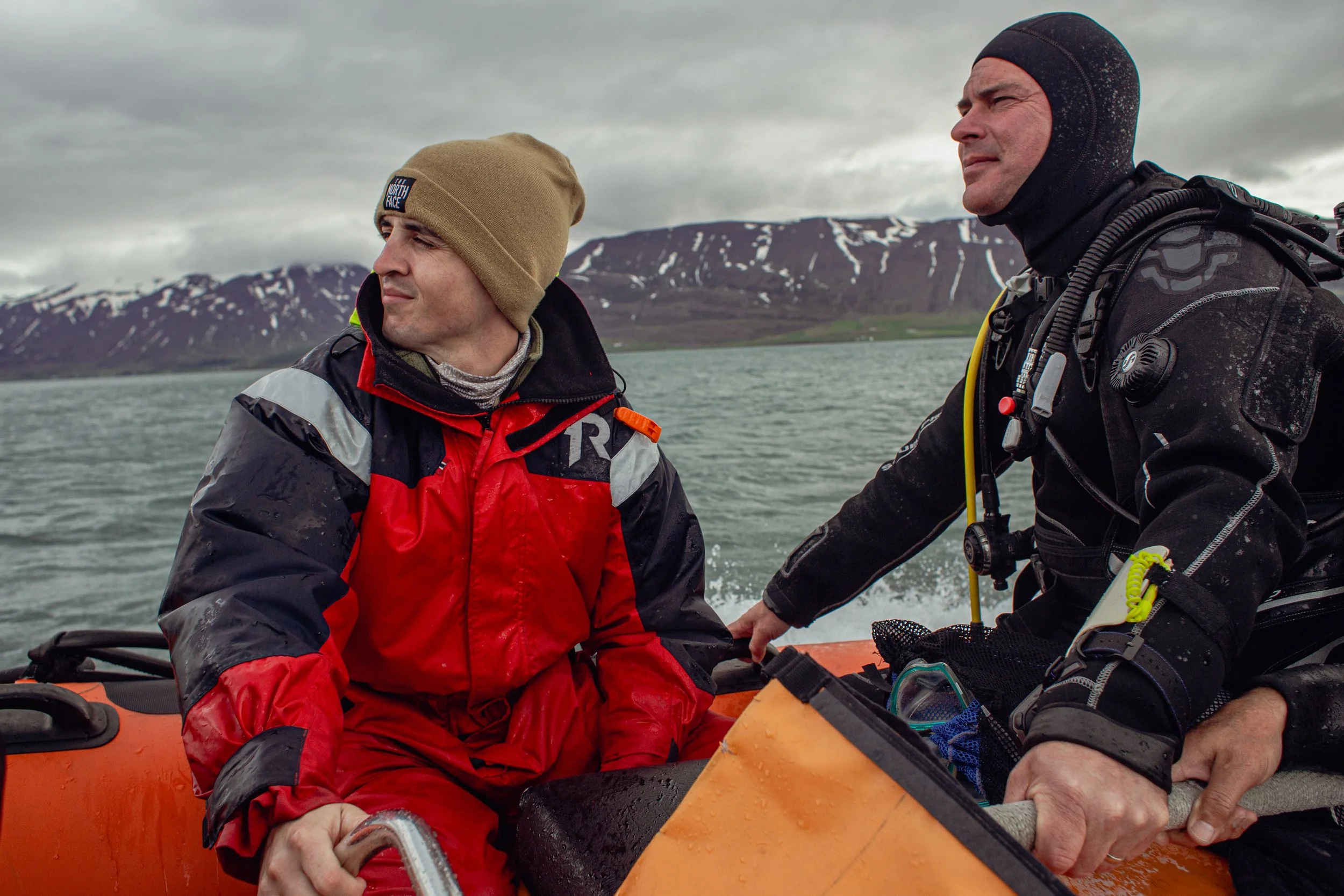 Two men in wetsuits and cold-weather gear on a rescue boat on a cold, cloudy day in a mountainous, snowy landscape.