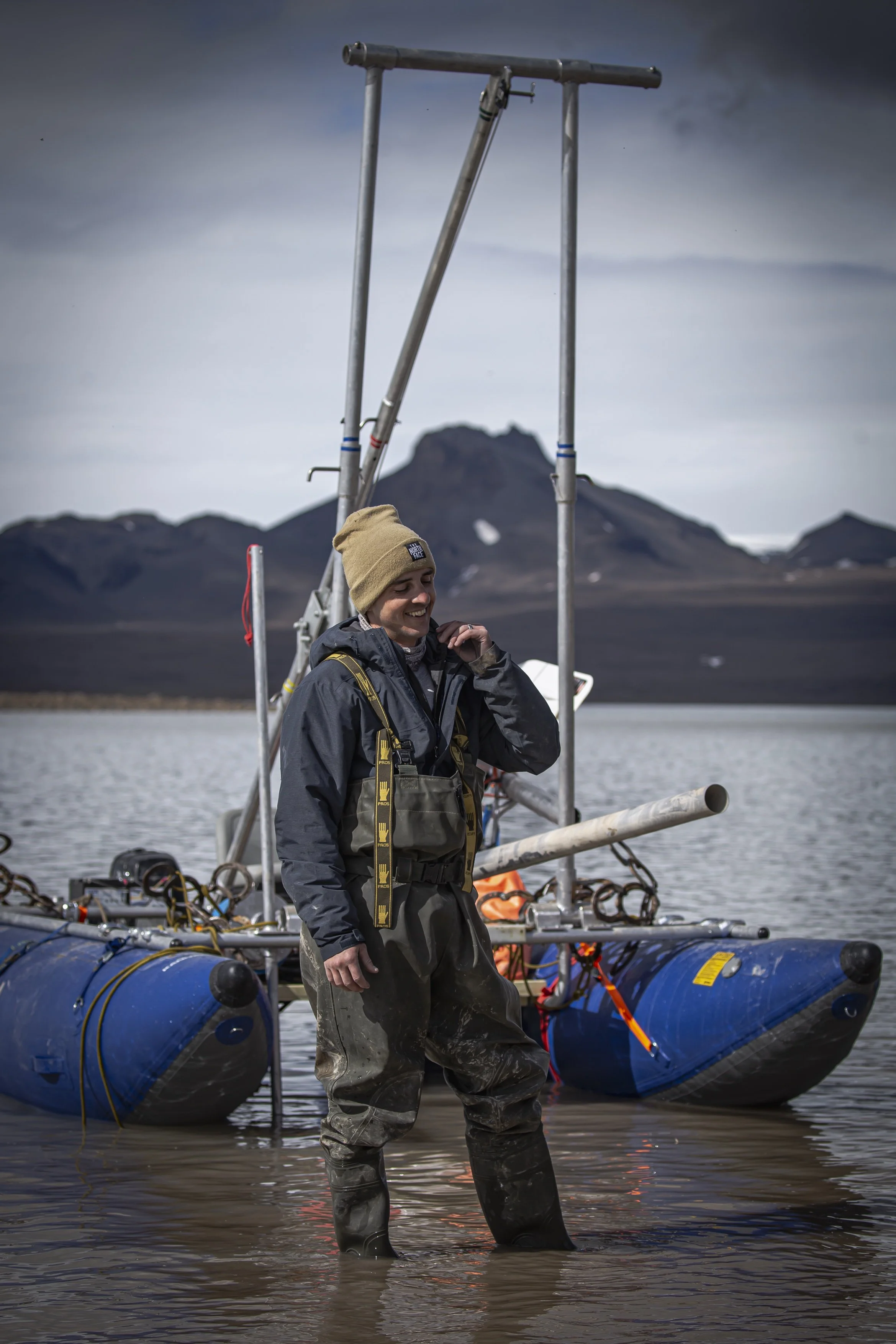 A person dressed in outdoor gear standing in shallow water near a blue hovercraft, with mountains and a cloudy sky in the background.