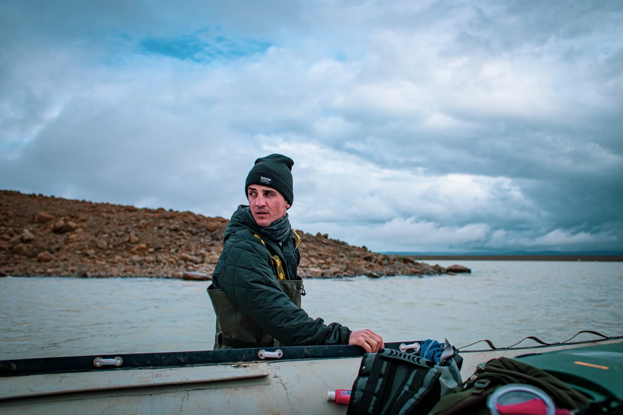 A young man in a black beanie, black jacket, and scarf stands in a boat on a body of water, with a rocky shoreline and cloudy sky in the background.