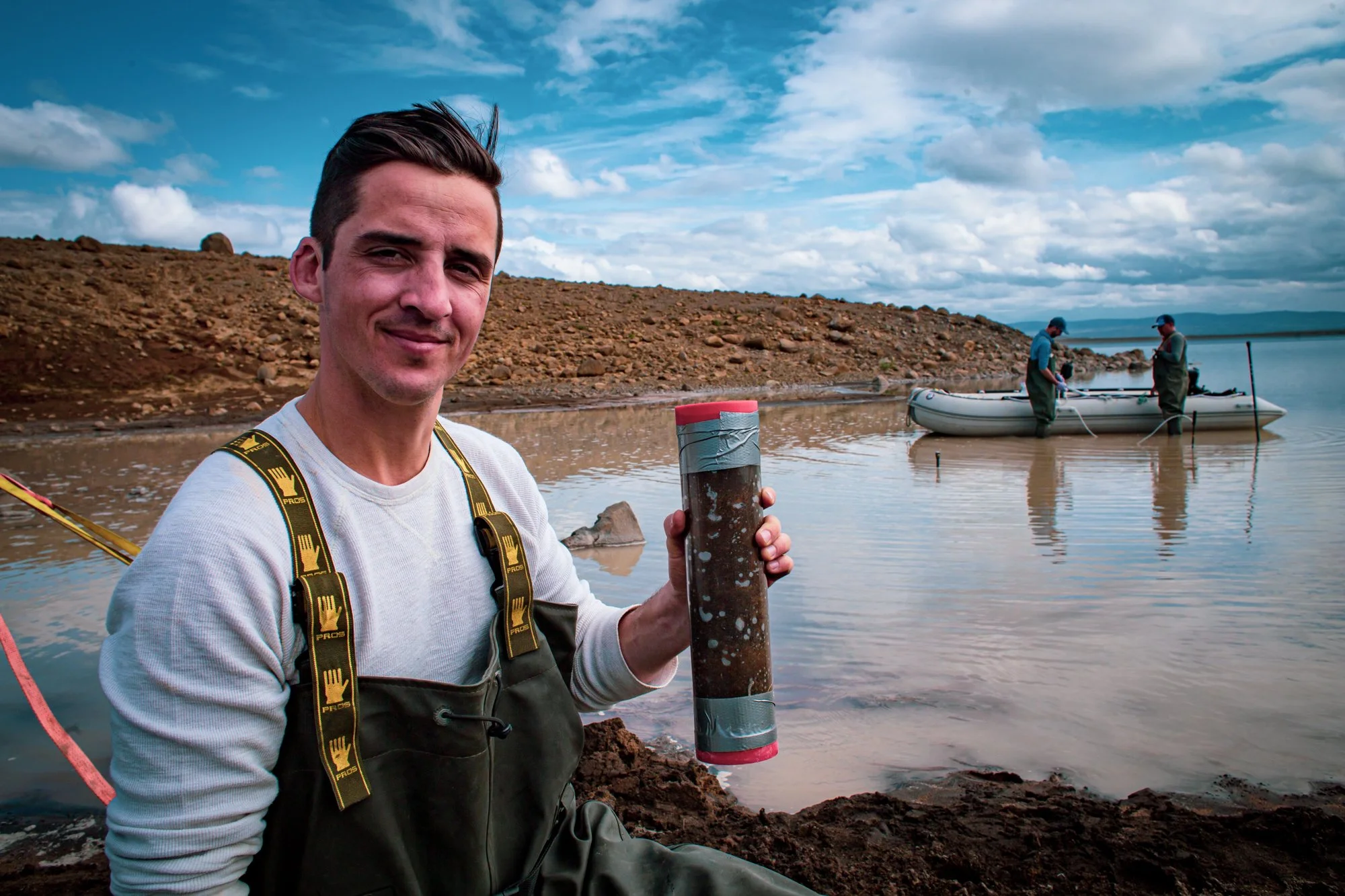 A man in fishing gear holding a cylindrical container near a body of water with two people in a small boat in the background and a rocky shoreline.