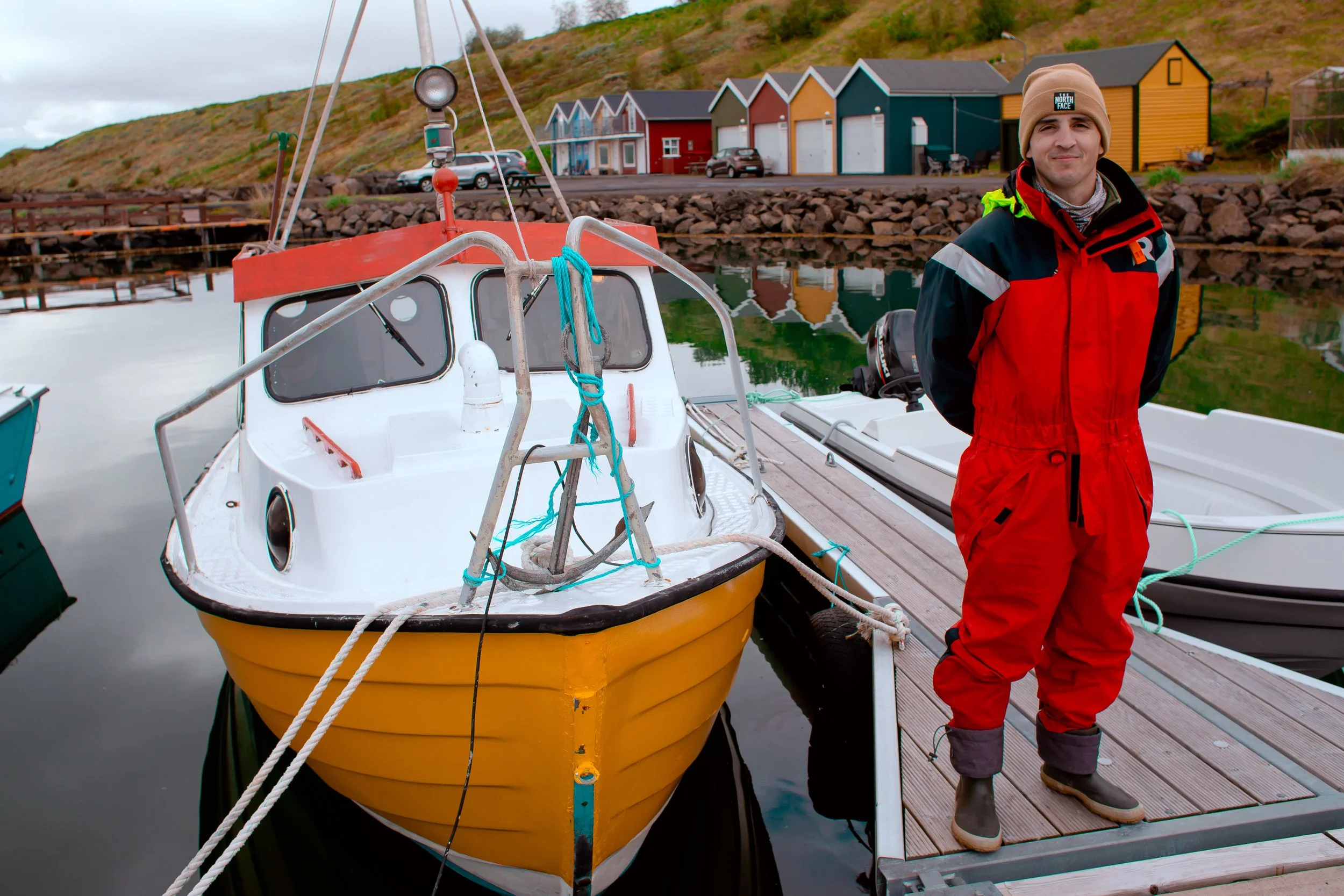A man in red and black waterproof gear standing on a wooden dock next to a yellow and white boat, with colorful small houses and hills in the background at a marina.