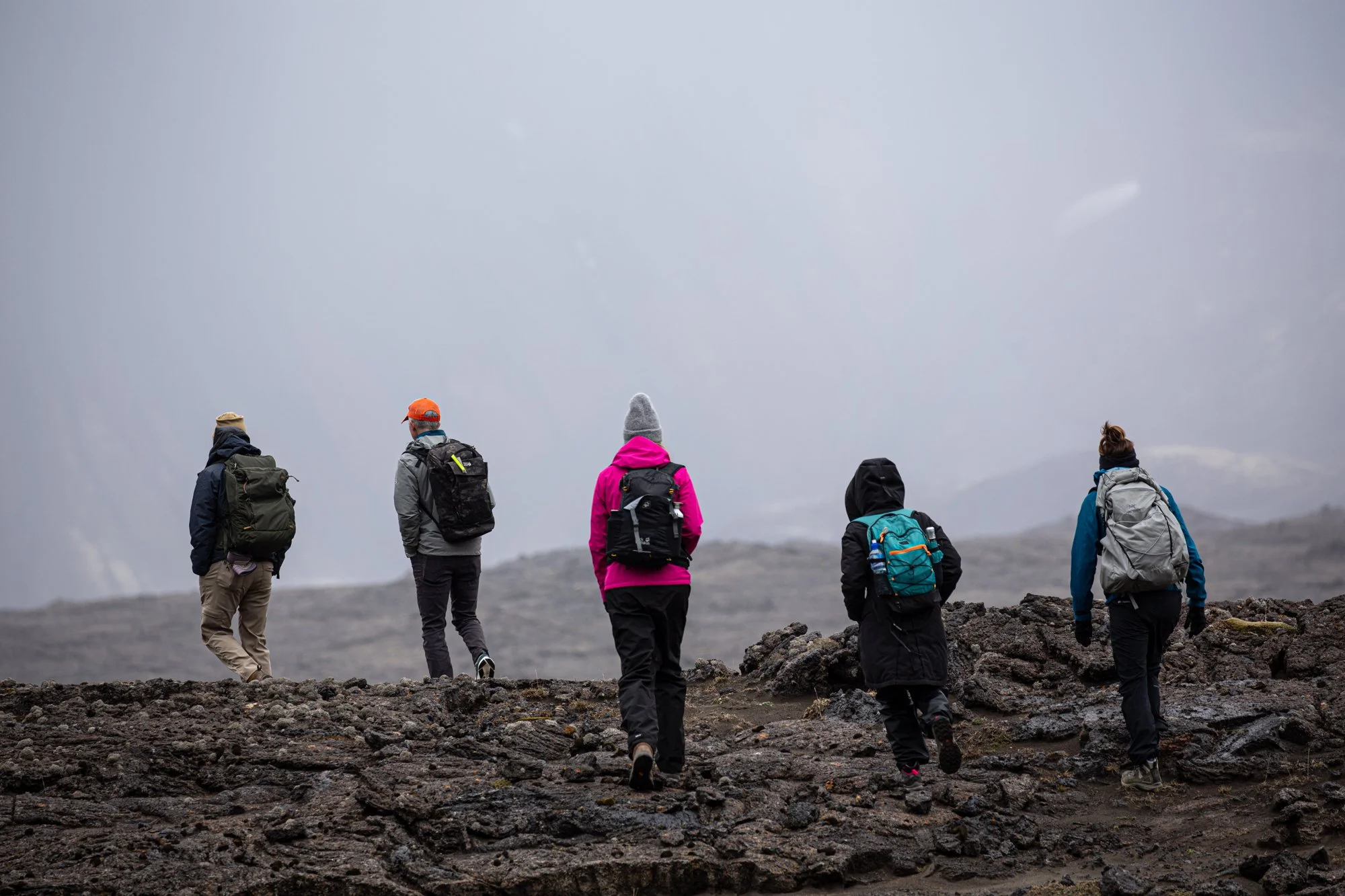 Five hikers with backpacks walking on rocky terrain in a cloudy, overcast landscape.