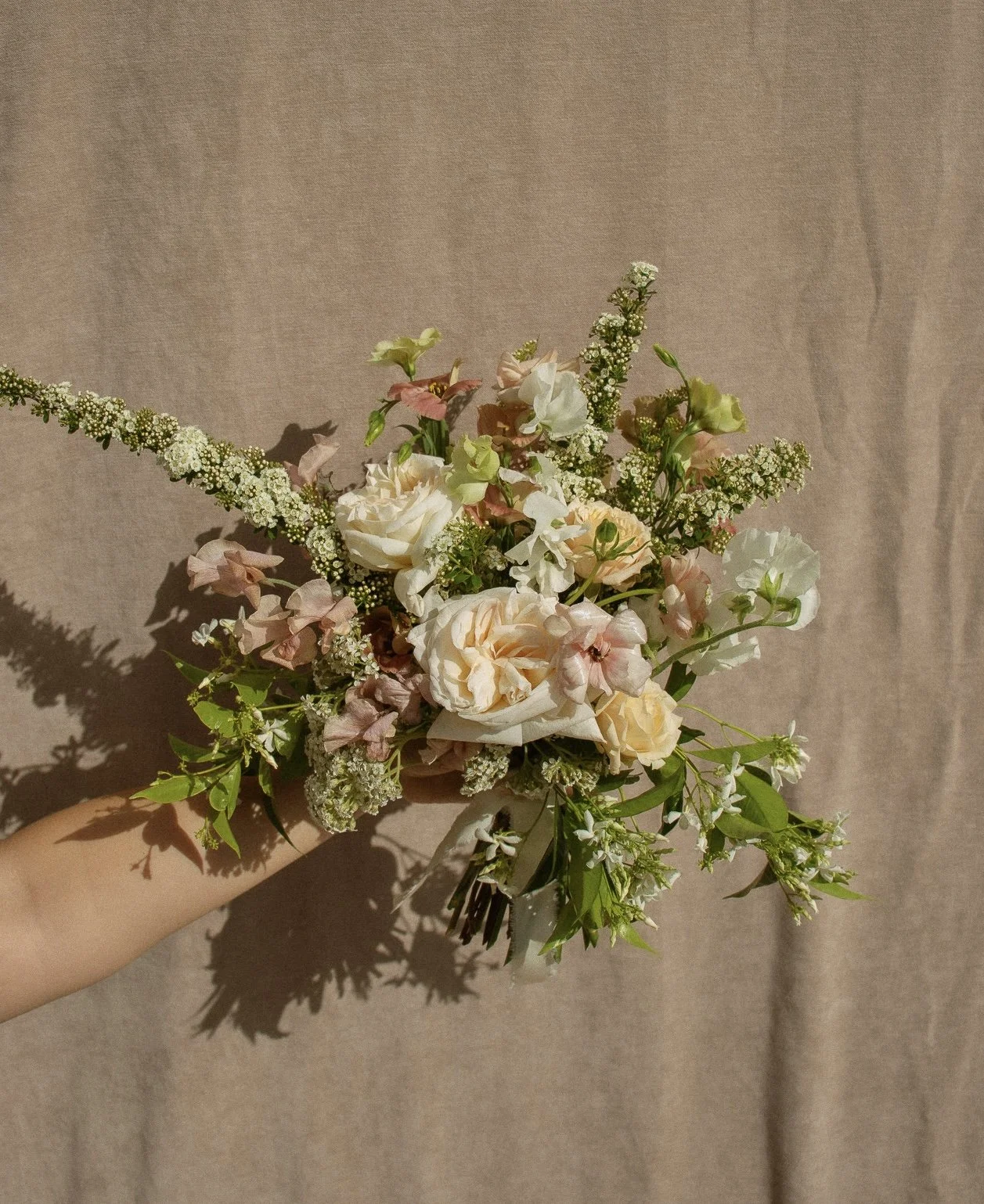 A hand holding a bouquet of mixed flowers including white roses, pink flowers, and greenery against a beige background.