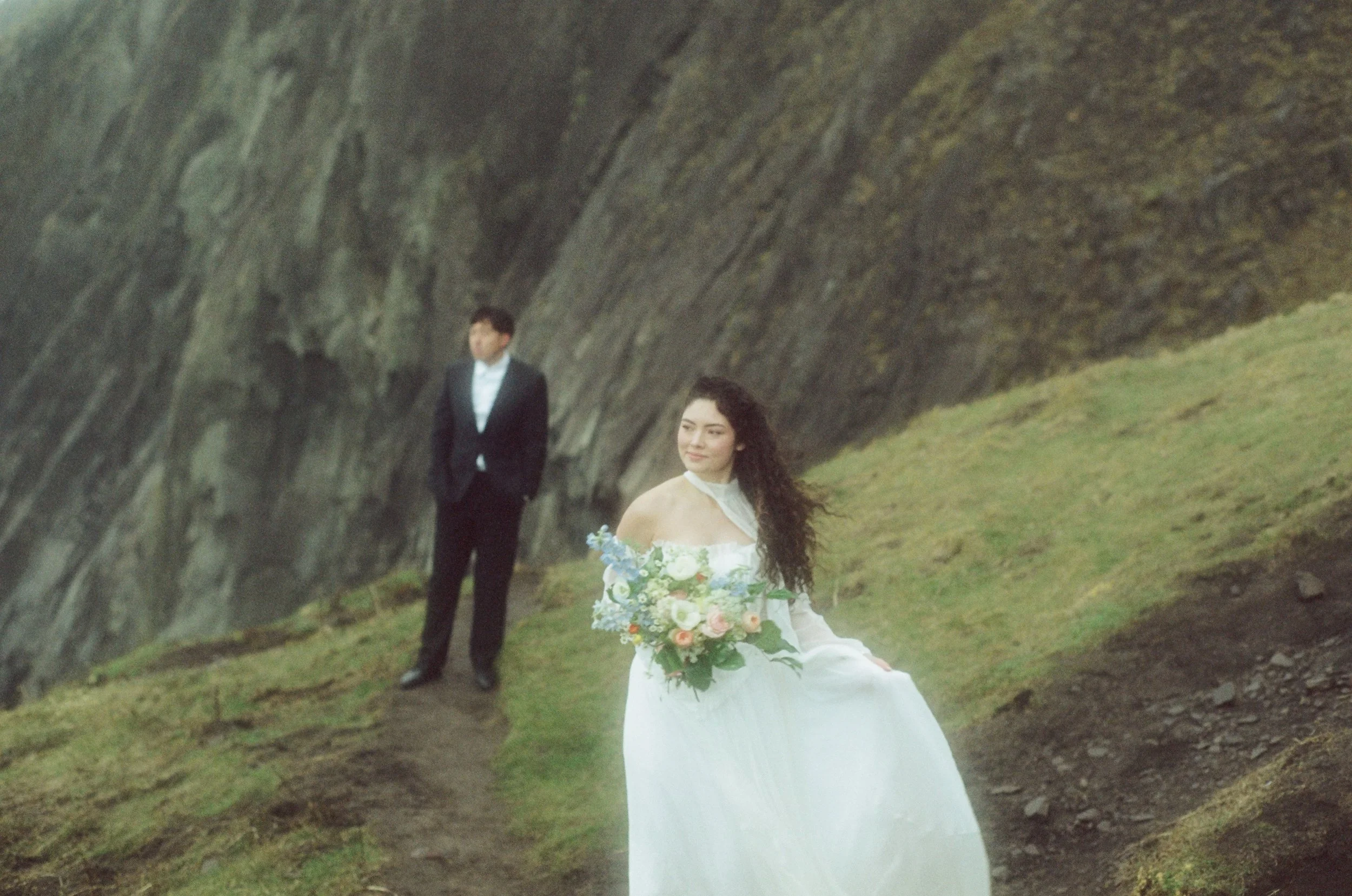 A woman in a wedding dress holding a bouquet of flowers standing on a grassy path with a man in a suit standing in the background near a rocky cliff.