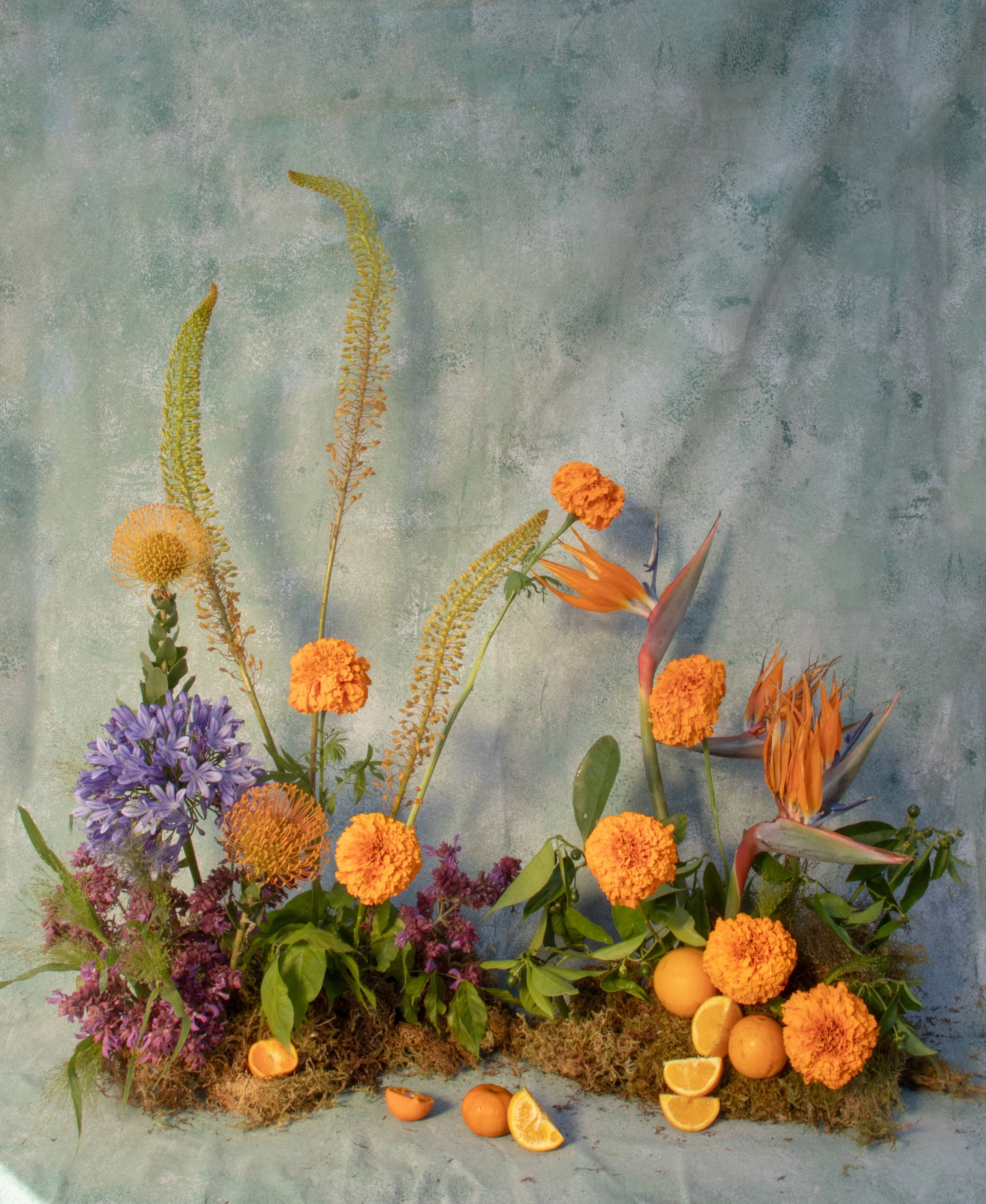 A floral arrangement with orange marigolds, purple flowers, orange birds of paradise, and green foliage, set against a textured blue-green background with oranges and orange slices at the base.