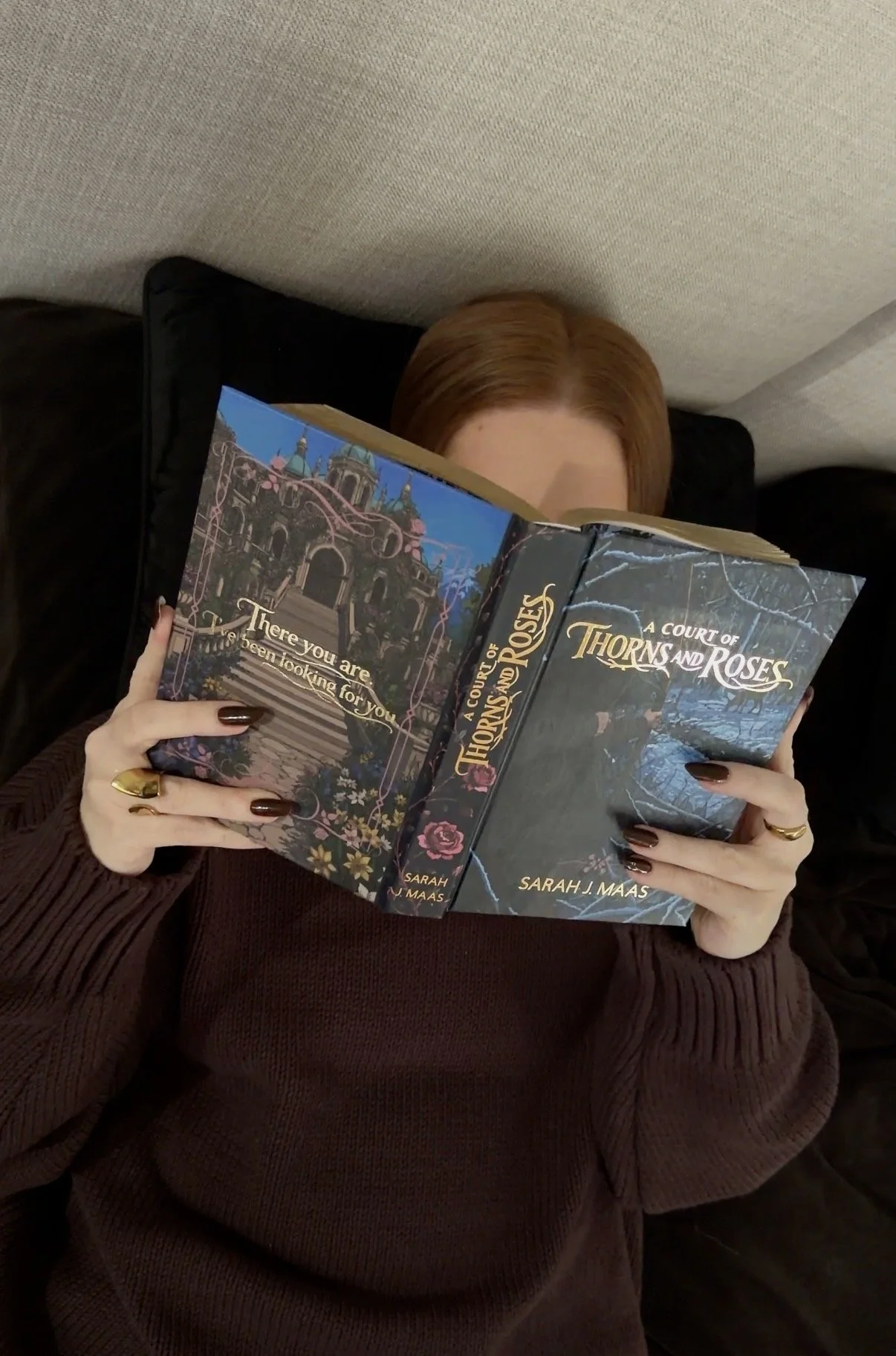 A person with red hair is lying on a couch and reading a book titled "A Court of Thorns and Roses" by Sarah J. Maas. The person's face is hidden behind the book, and they are wearing a dark red sweater with matching nail polish and gold rings.