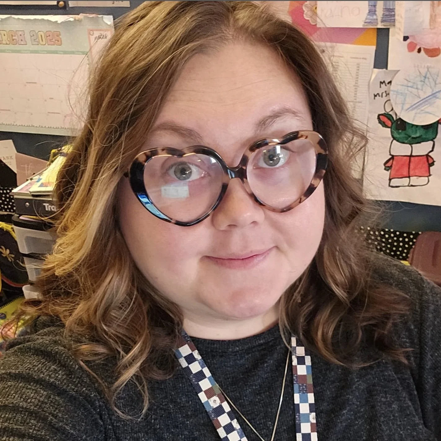 A woman with curly hair wearing tortoise shell heart-shaped glasses, a dark sweater, and a checkered lanyard, taking a selfie in a colorful, decorated room.
