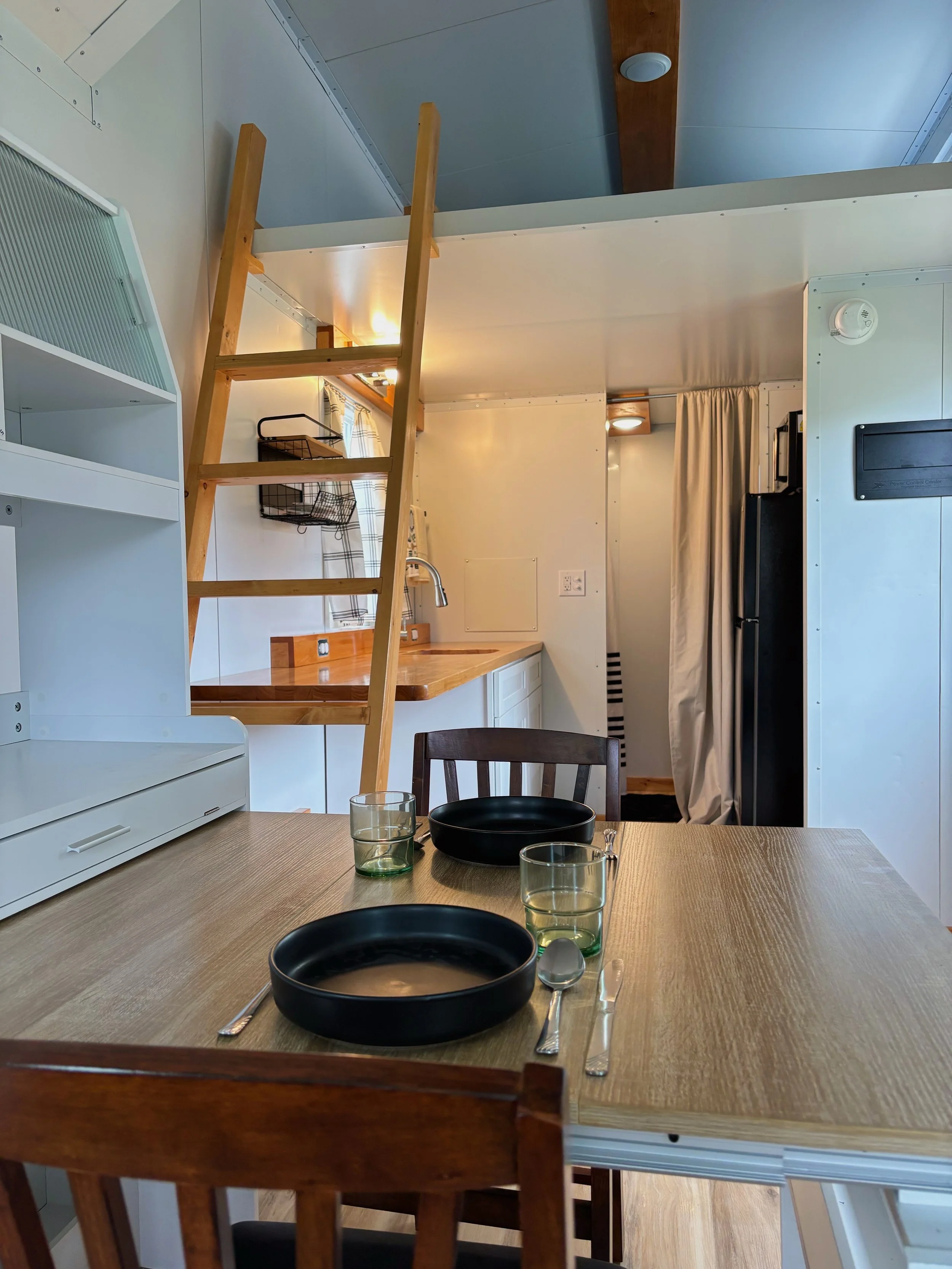 Inside a tiny home or apartment with a wooden dining table set with black plates, glasses, and utensils. In the background, a small kitchen area with a sink, countertop, refrigerator, and a loft with a wooden ladder leading up to it.