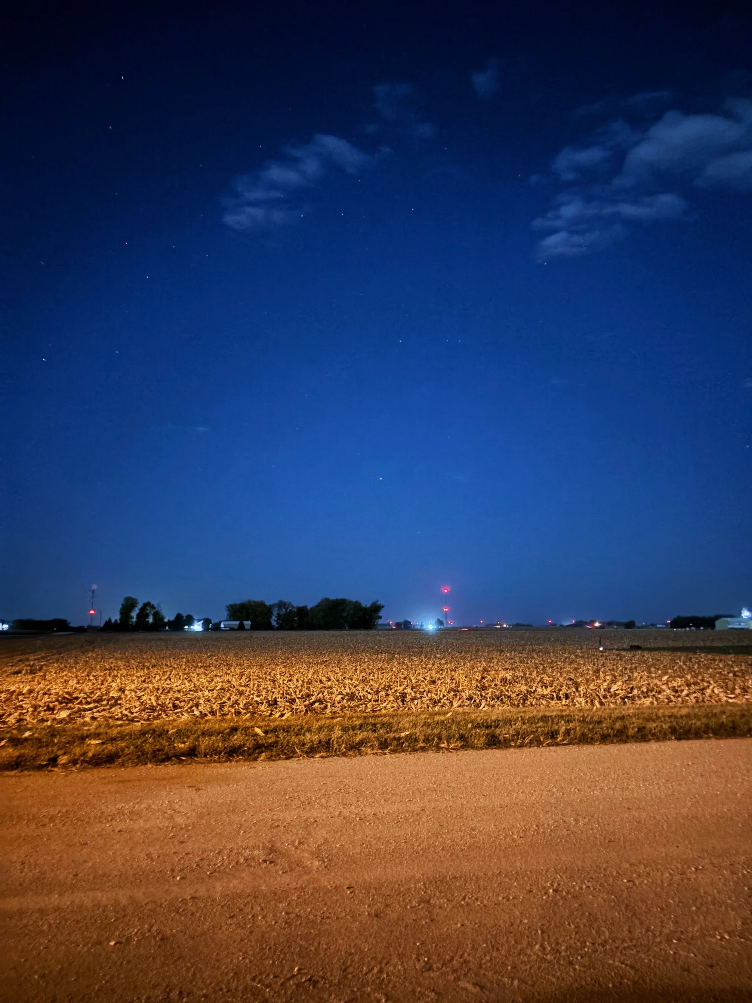 Night sky with stars, clouds, a large field in the foreground, and distant trees and communication towers with red lights.
