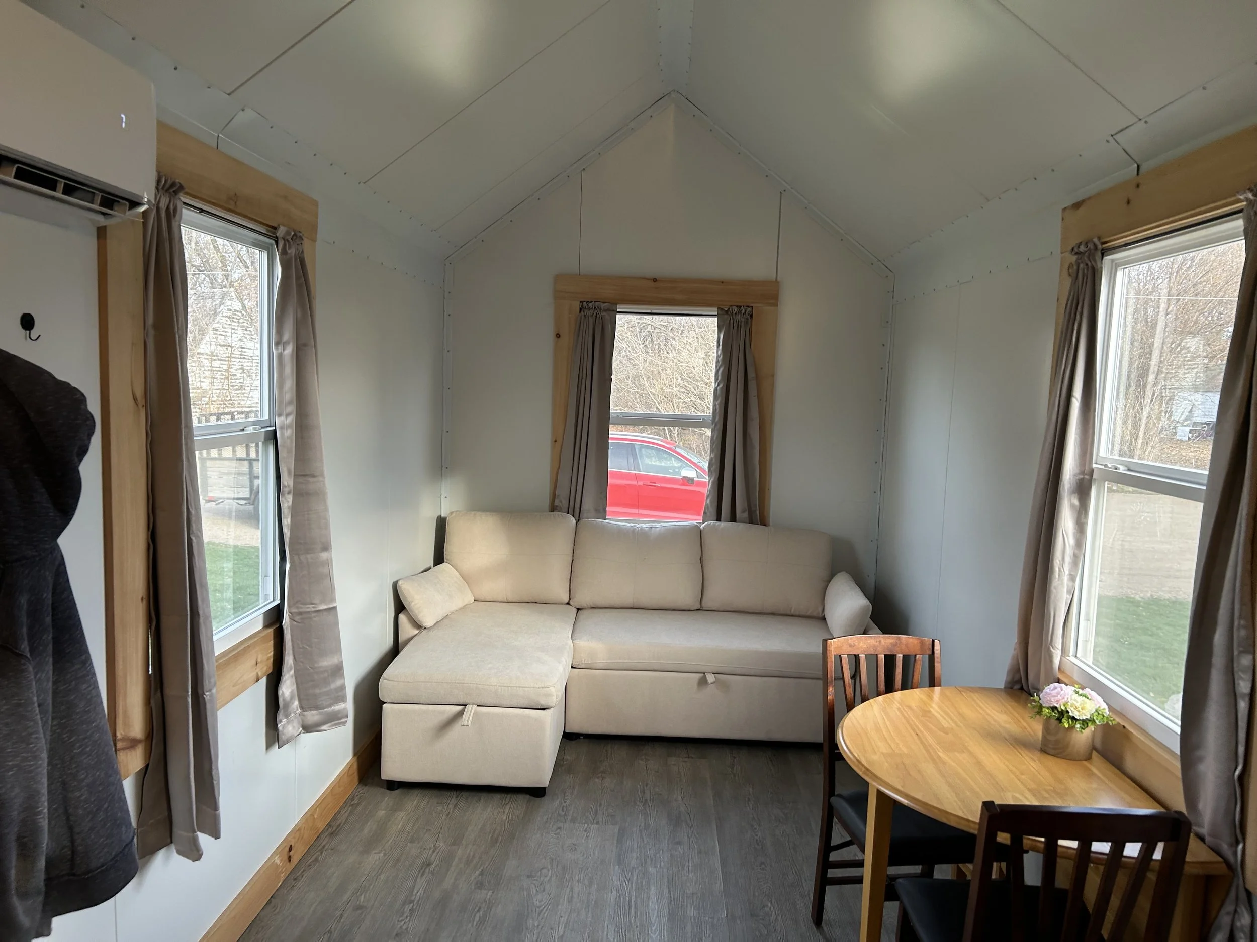 Small living room with a beige sectional sofa, wooden framed windows with beige curtains, a round wooden table with two chairs, and a small flower arrangement on the table.