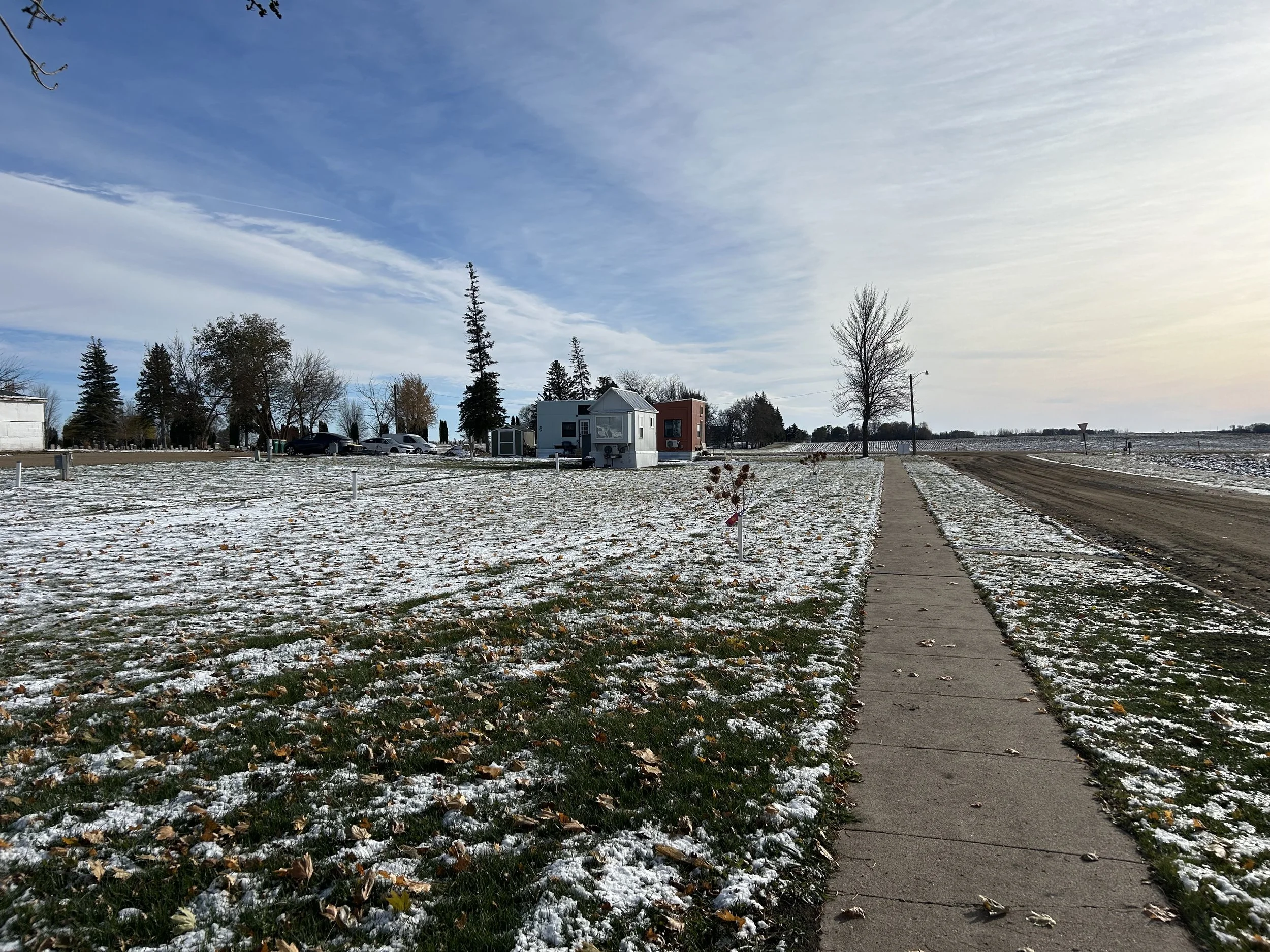 A sidewalk leading towards small residential buildings in a snow-covered open area on a cloudy day.