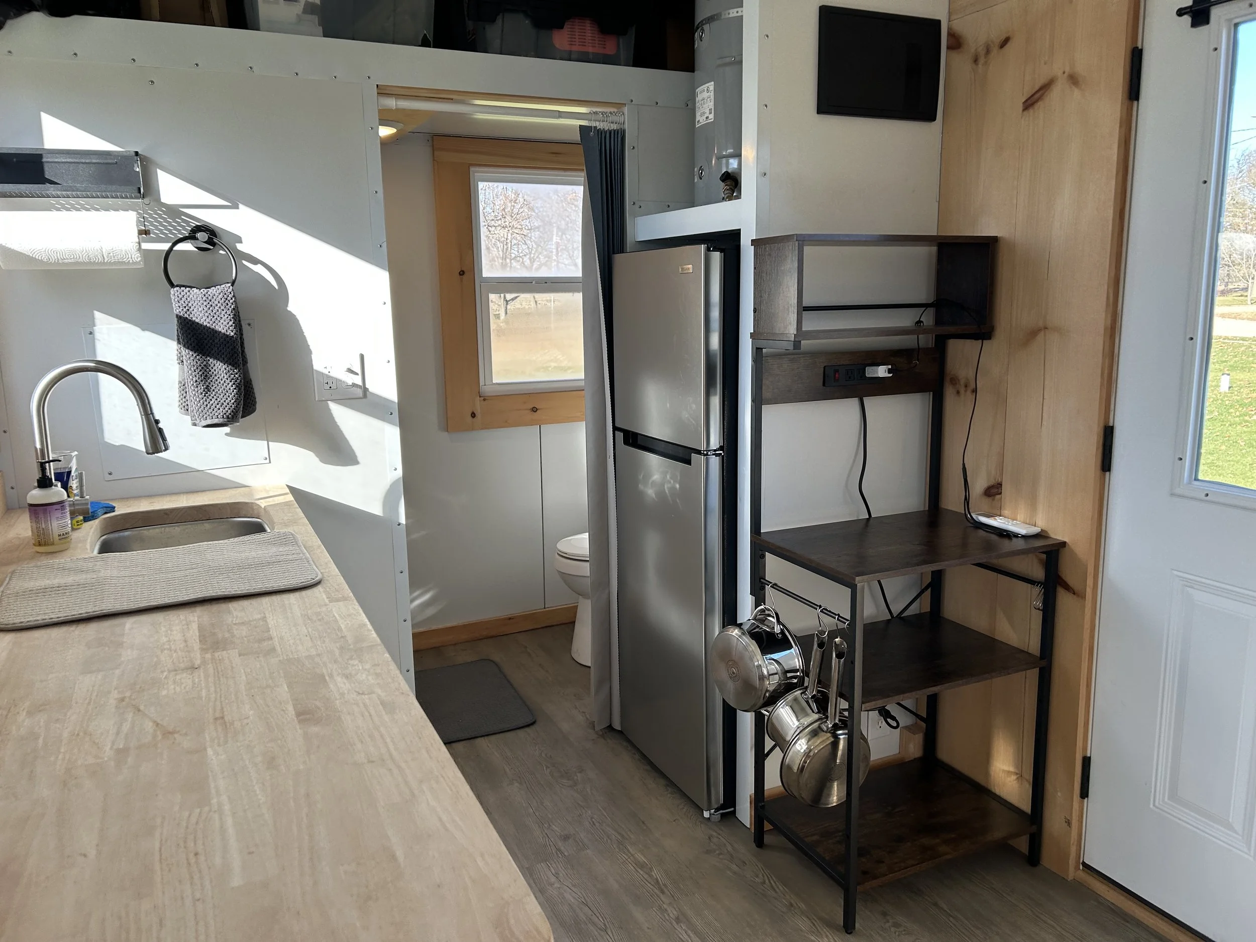 Interior of a small kitchen with a wooden countertop, a stainless steel sink, a small refrigerator, a wooden shelf unit, and a window with a view of an outdoor landscape. There's a towel hanging on a hook near the sink.