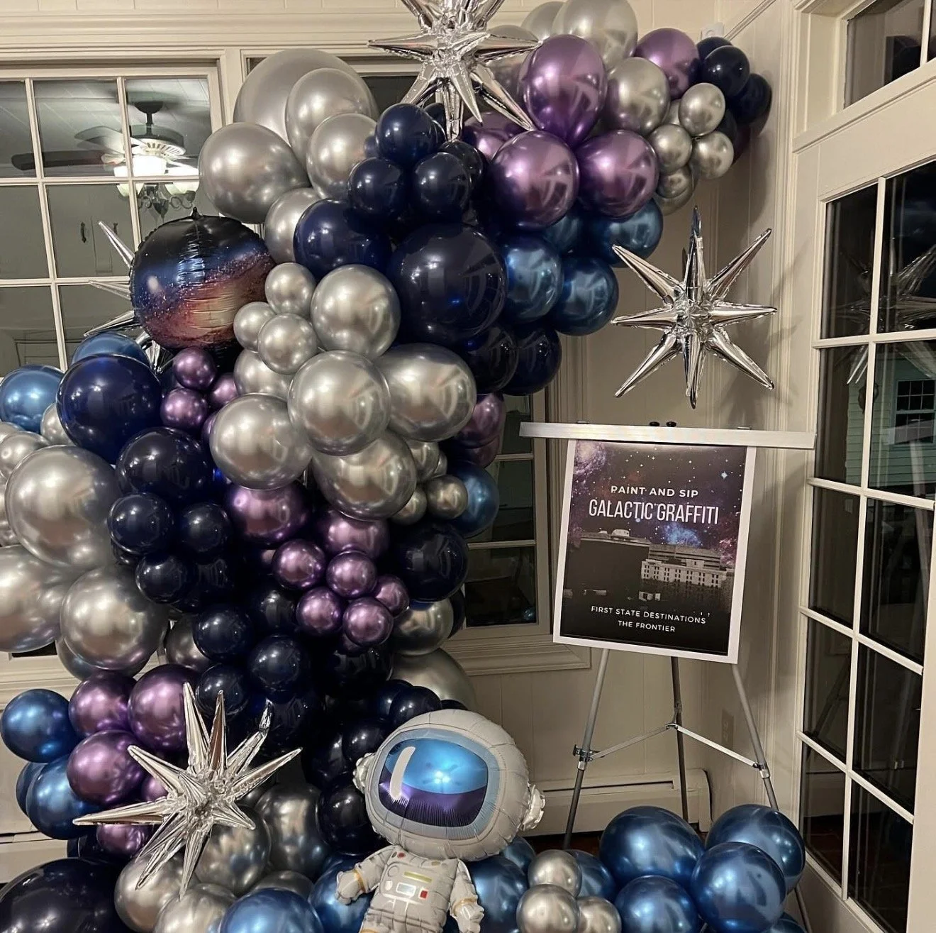 Custom balloon arch installation of blue, purple, silver balloons with an astronaut balloon and stars in an office space by The Design Ward for a paint and sip event
