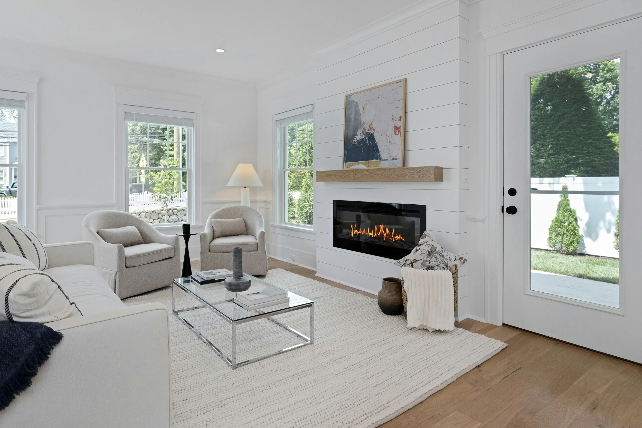 Bright living room with white walls, multiple windows, a white sofa with pillows, a glass coffee table with books and decor, a white armchair, a coat of arms fireplace, and large windows showing green trees outside.