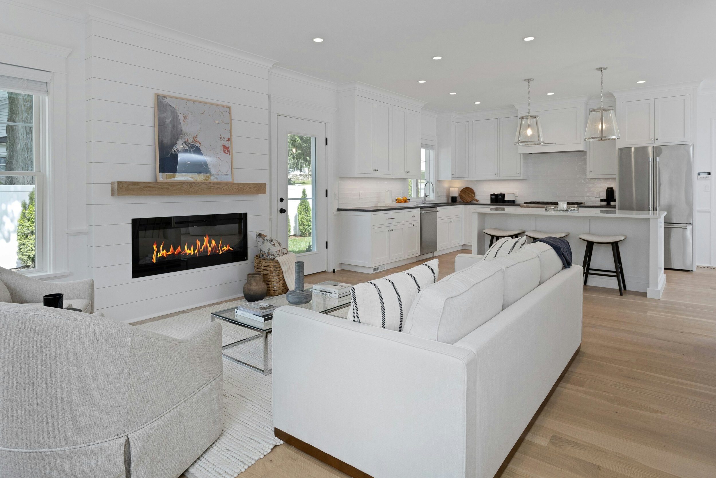 Bright living room with white furniture, a fireplace, and an open kitchen in the background.