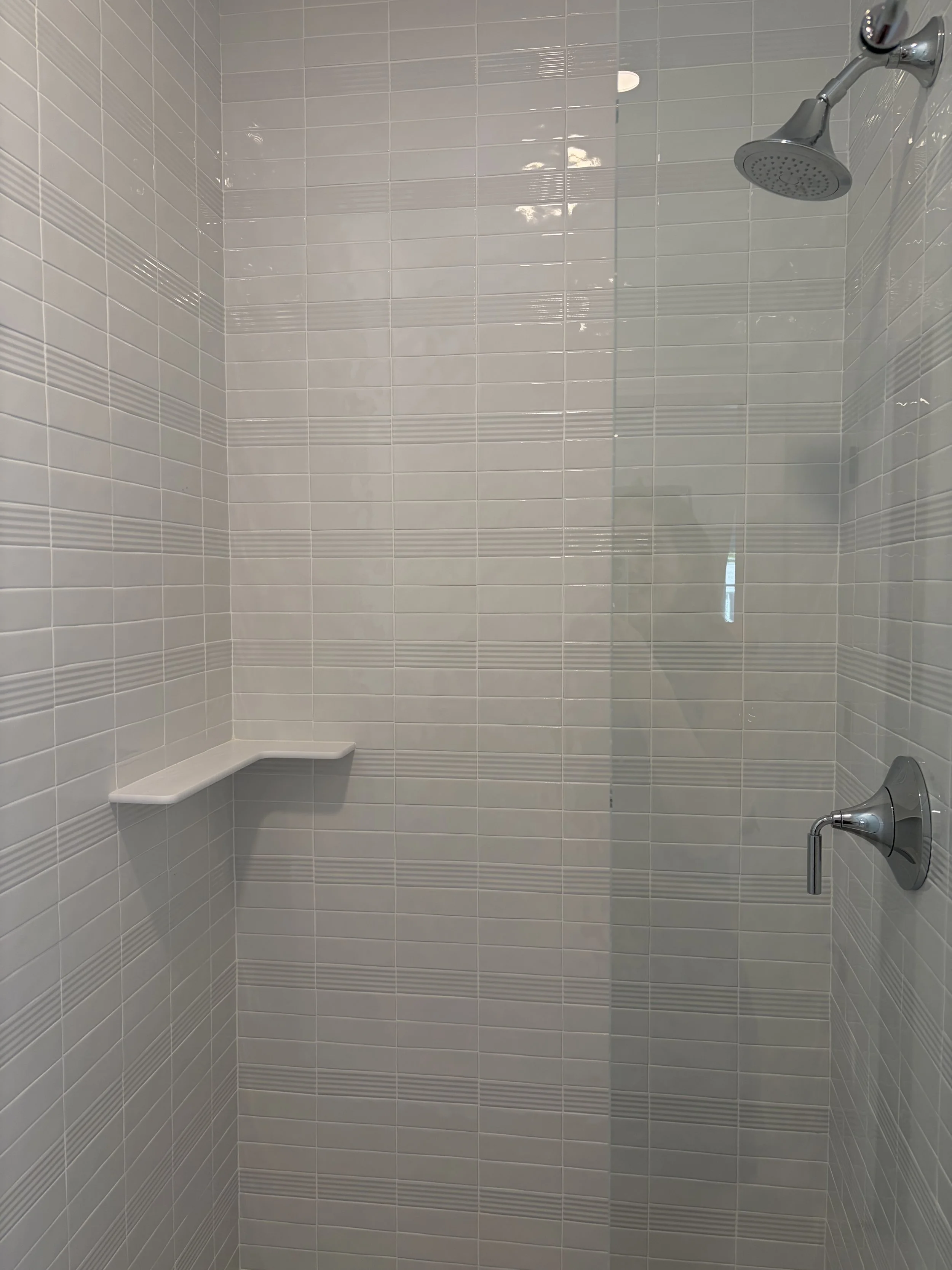 Shower area with white tiles, a showerhead, a handheld shower attached on the right, and a corner shelf on the lower-left side.