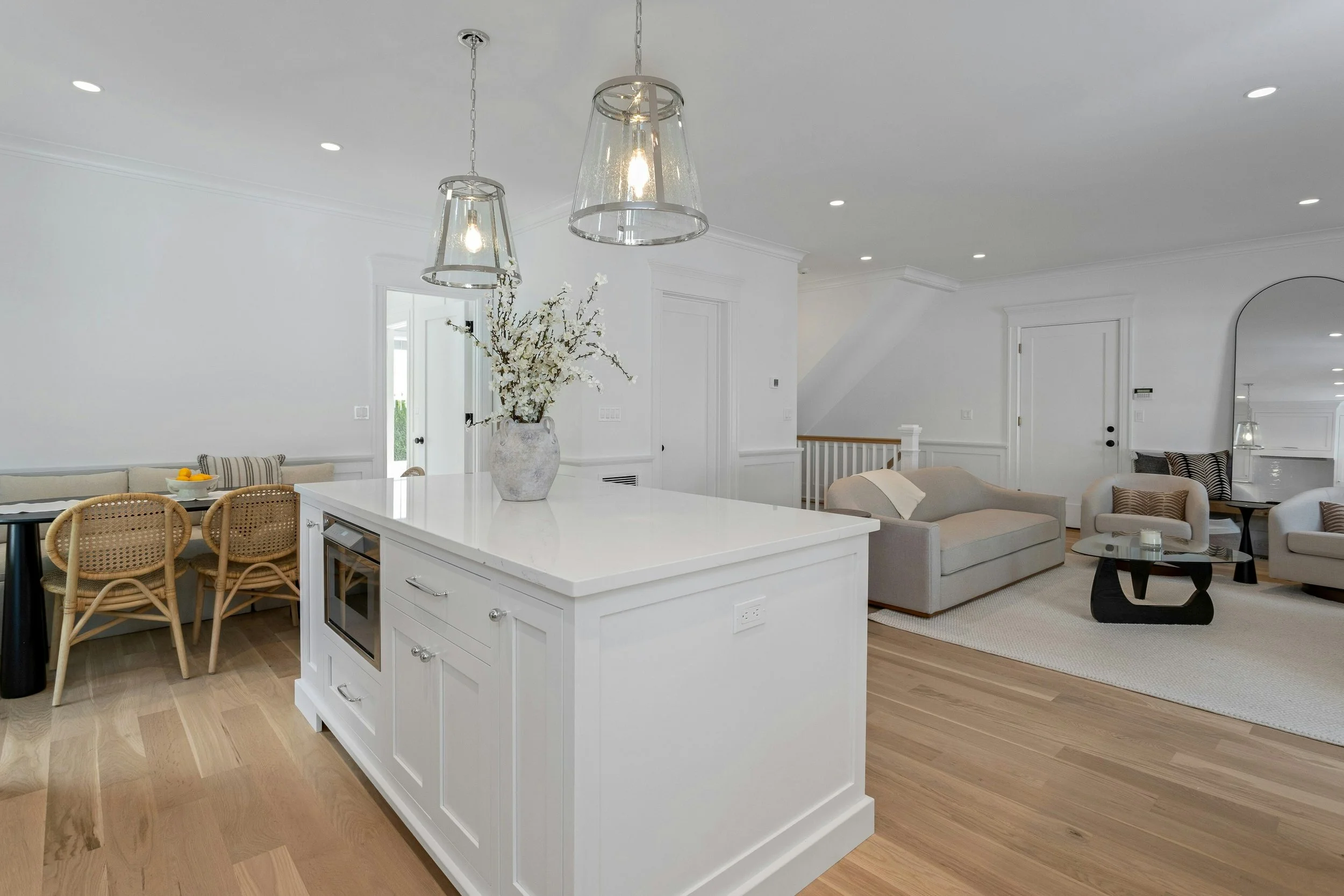 Open-concept living room and kitchen with white walls, hardwood floors, and modern furniture. There are two pendant lights hanging from the ceiling, a large floral vase on the kitchen island, and seating including chairs and sofas.