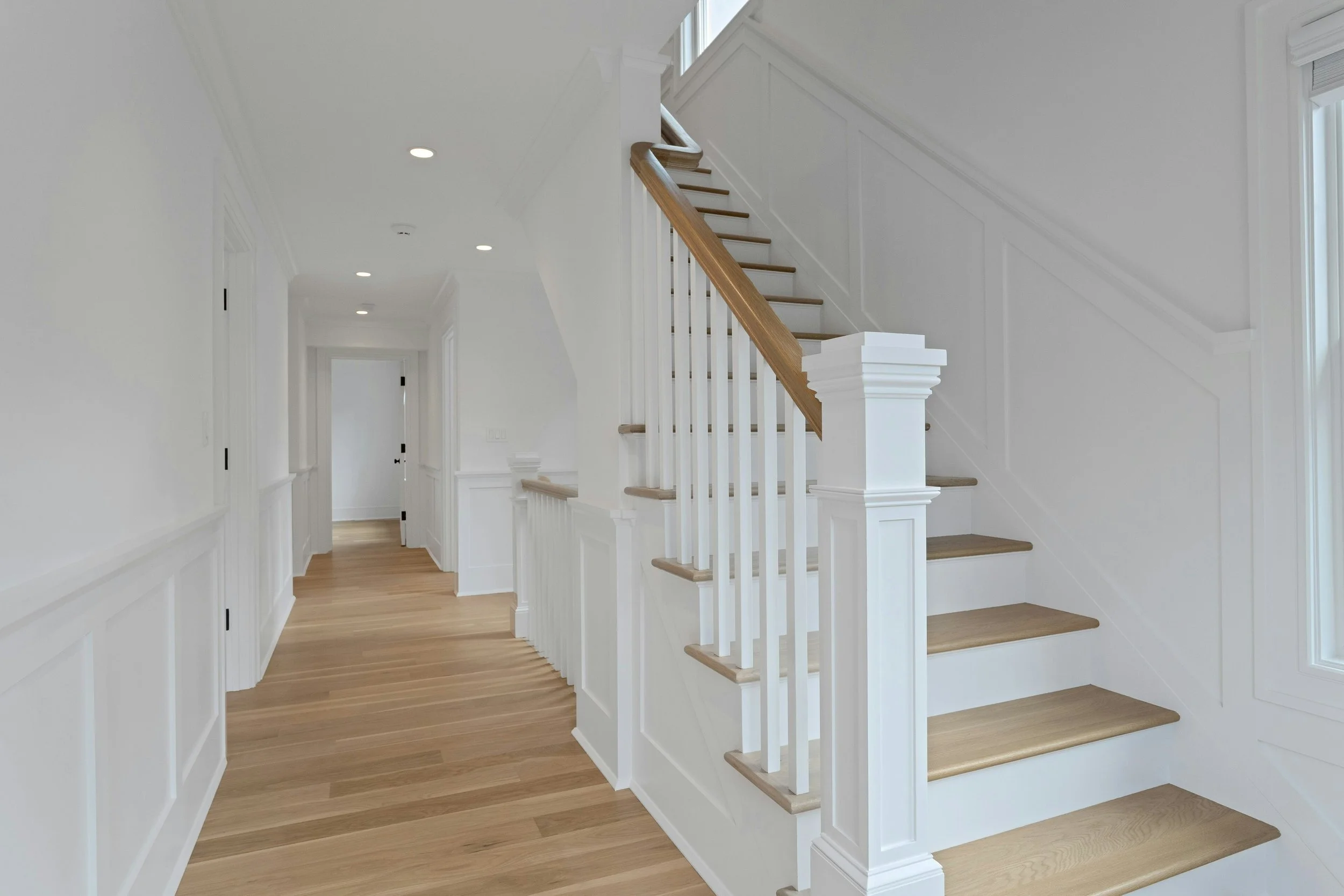 Interior view of a bright, white staircase hallway with wooden floors, white walls, and a staircase with wooden handrails and white balusters leading upward. Multiple round ceiling lights illuminate the space.