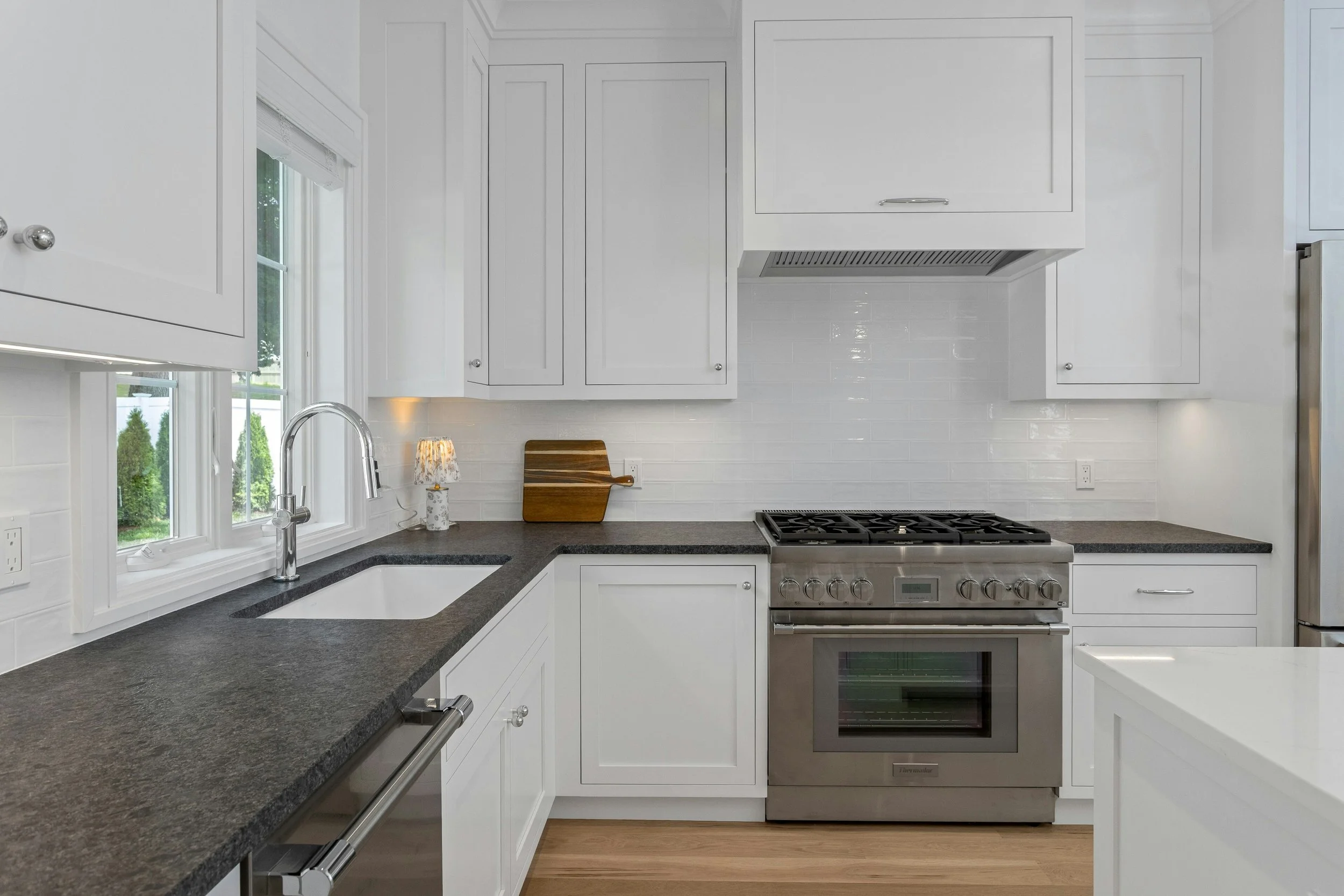 A modern kitchen with white cabinets, black countertops, a stainless steel oven, and a window above the sink.
