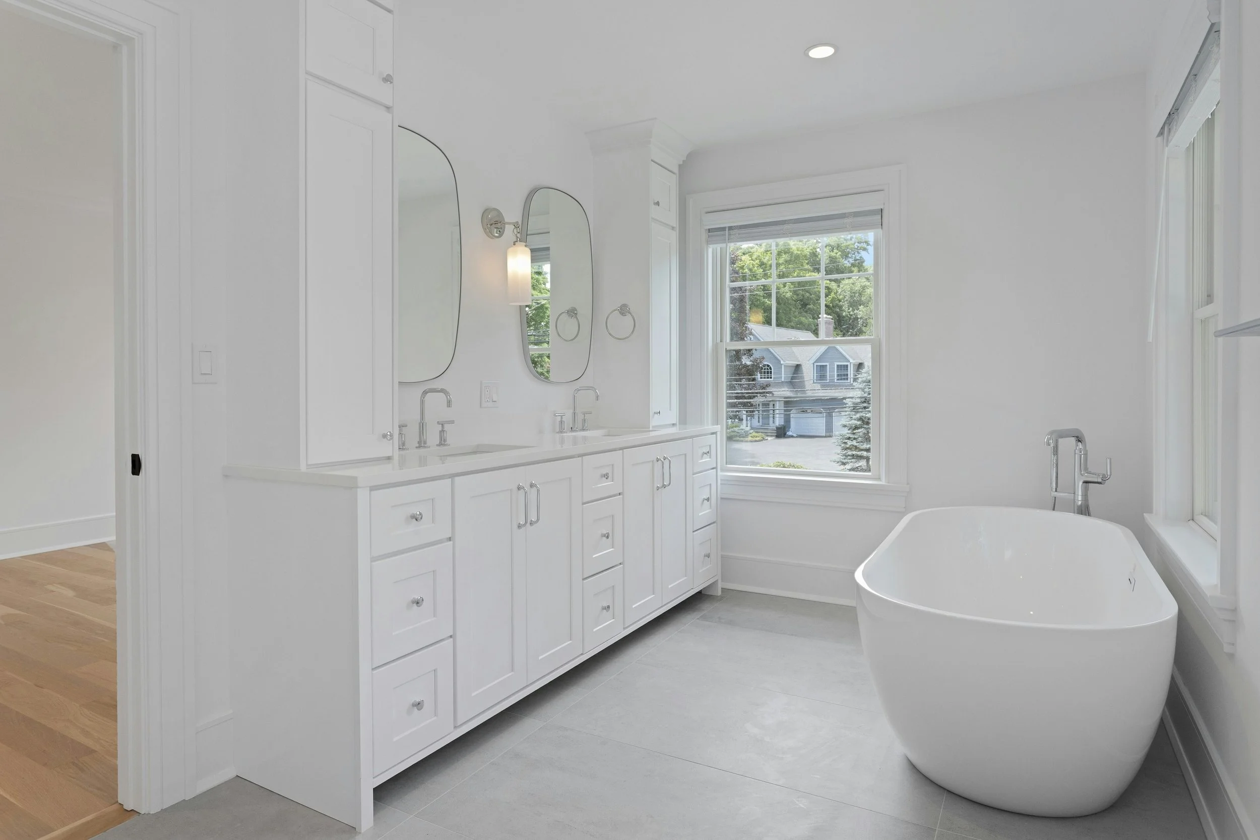 A bright, modern bathroom with a large window showing a neighborhood view, featuring a white vanity with two mirrors, a white bathtub, and white walls.