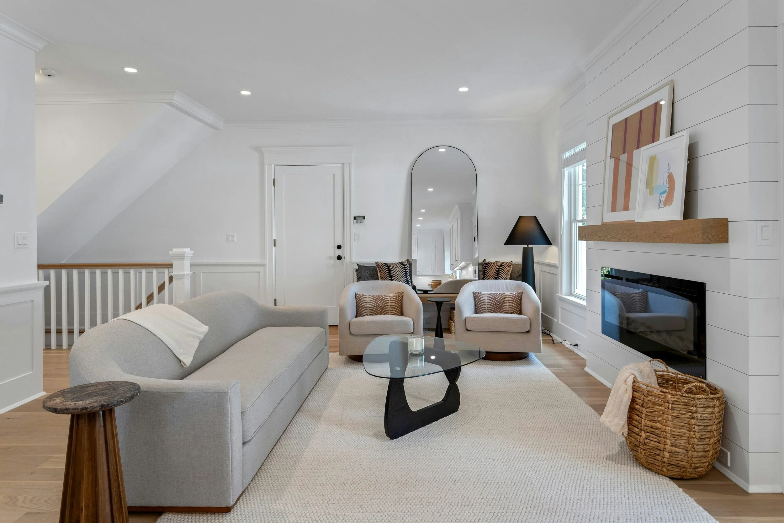 Living room with white walls, a beige couch, matching armchairs, a black coffee table, a black floor lamp, artwork leaning against the wall, a wooden side table, a basket, and a fireplace, with natural light coming through the window.