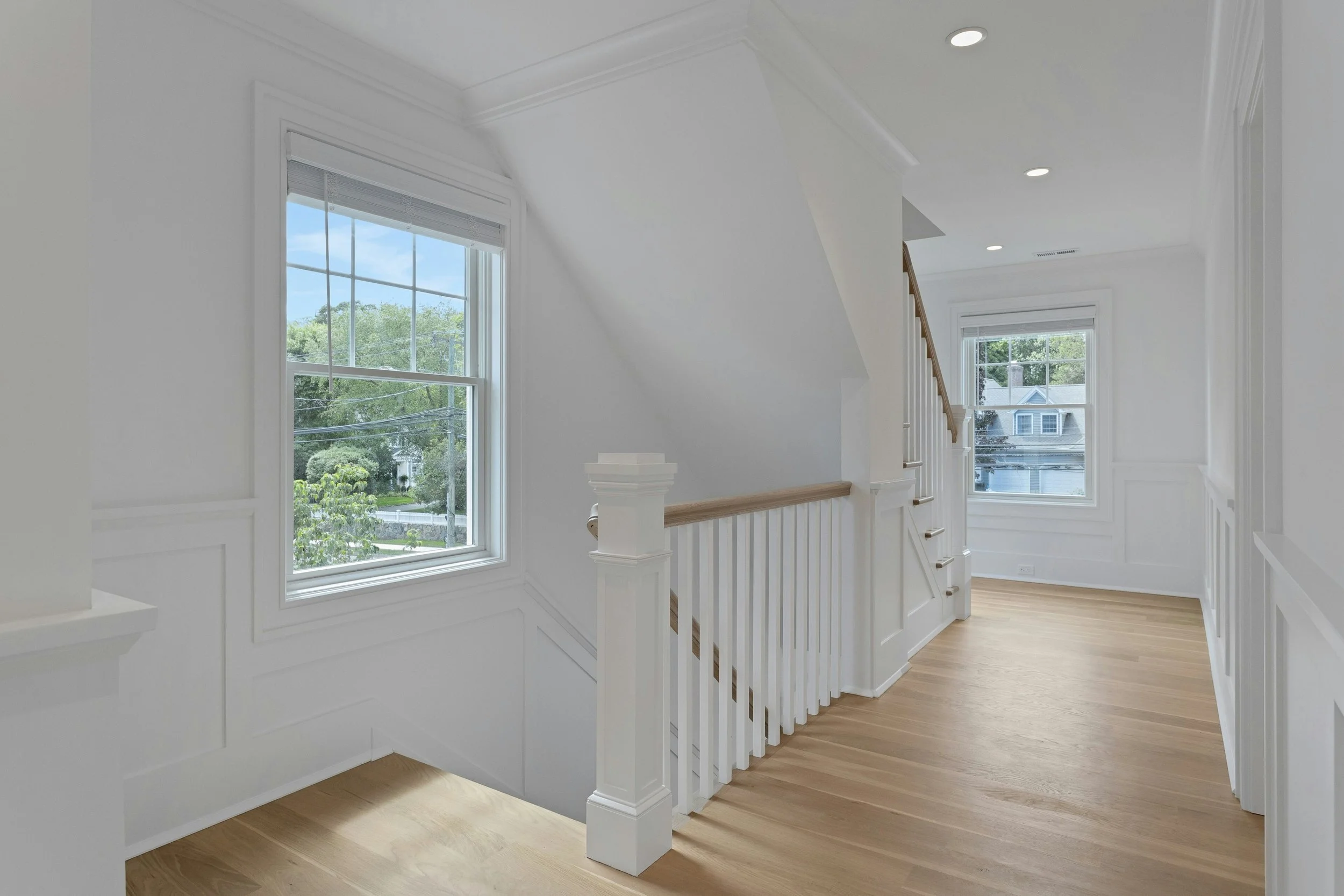 Interior view of a bright stairway landing with large windows, white walls, wooden floors, and white wooden banisters.