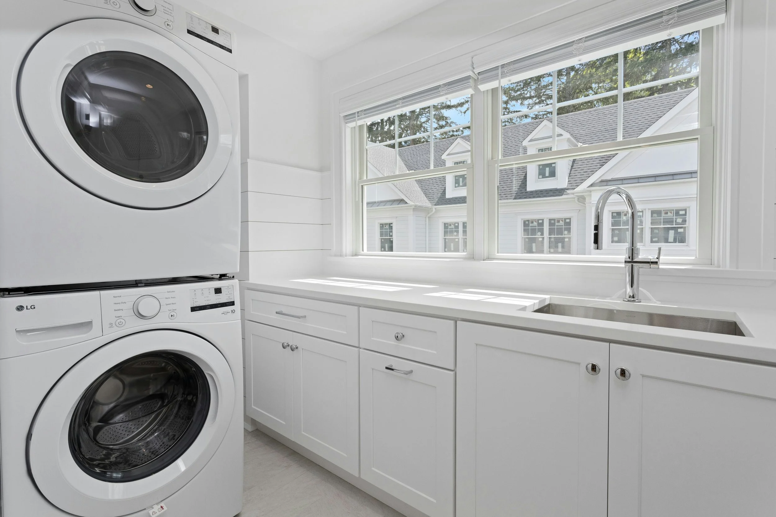 A laundry room with stacked white washer and dryer, white cabinets, a large window showing a neighborhood with houses, and a sink with a chrome faucet.