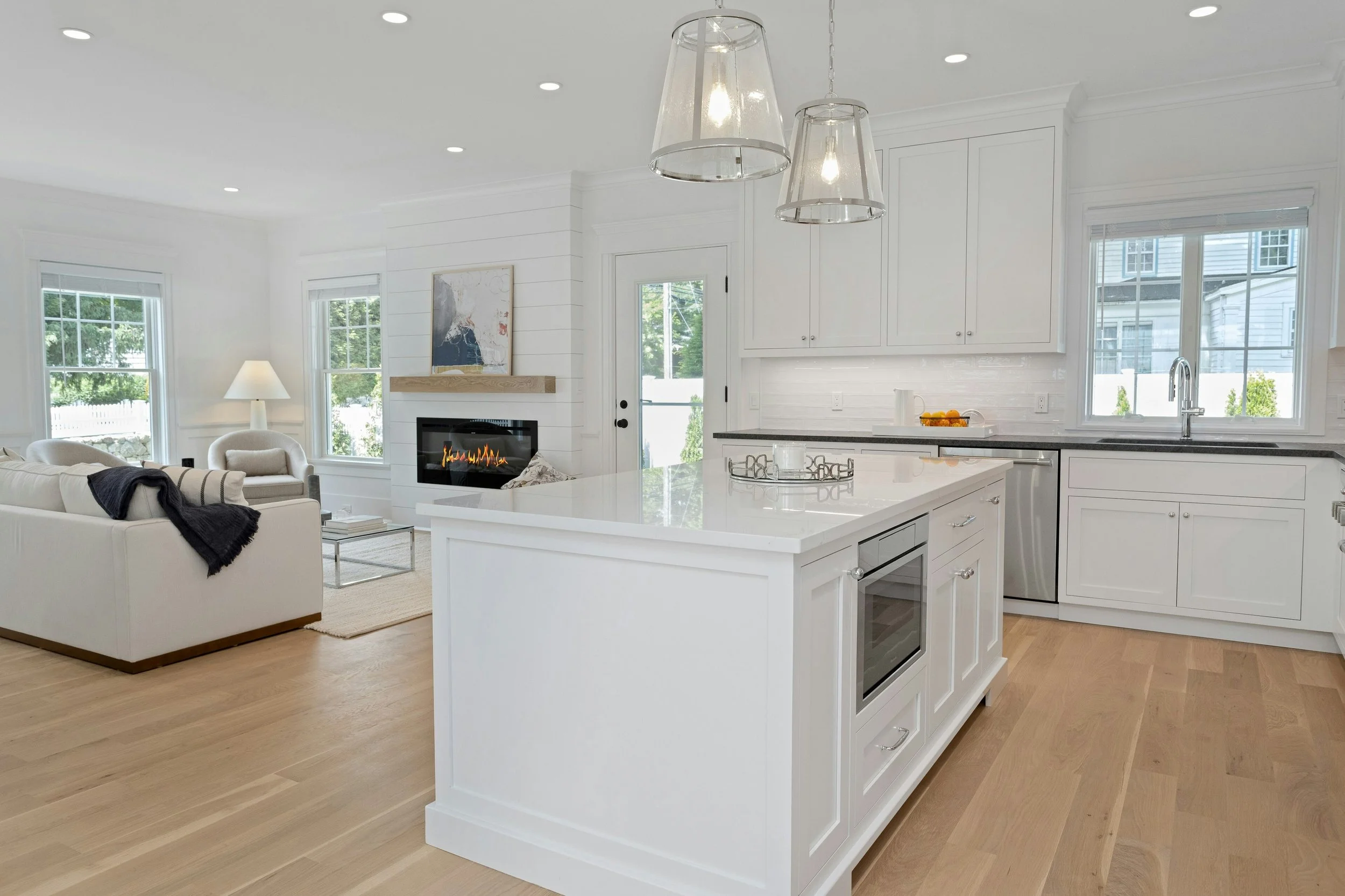 Bright, modern open-concept kitchen and living room with white cabinetry, large windows, a fireplace, and minimalistic furniture.