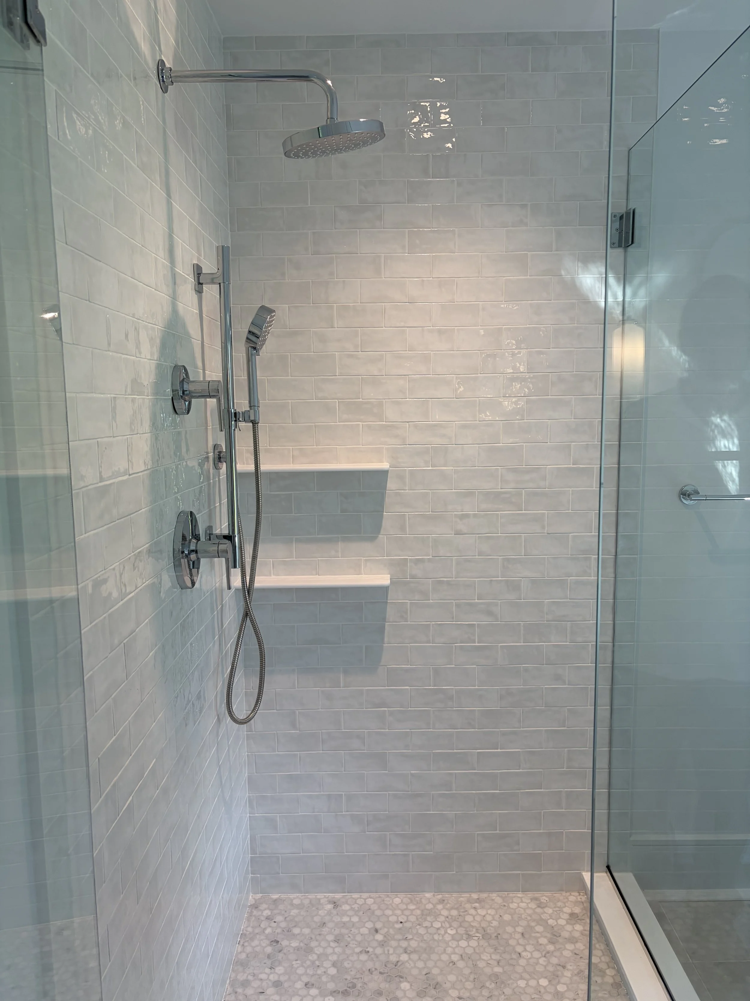 Modern shower area with chrome showerhead and handheld sprayer, tiled with subway tiles, and a partially visible glass enclosure.