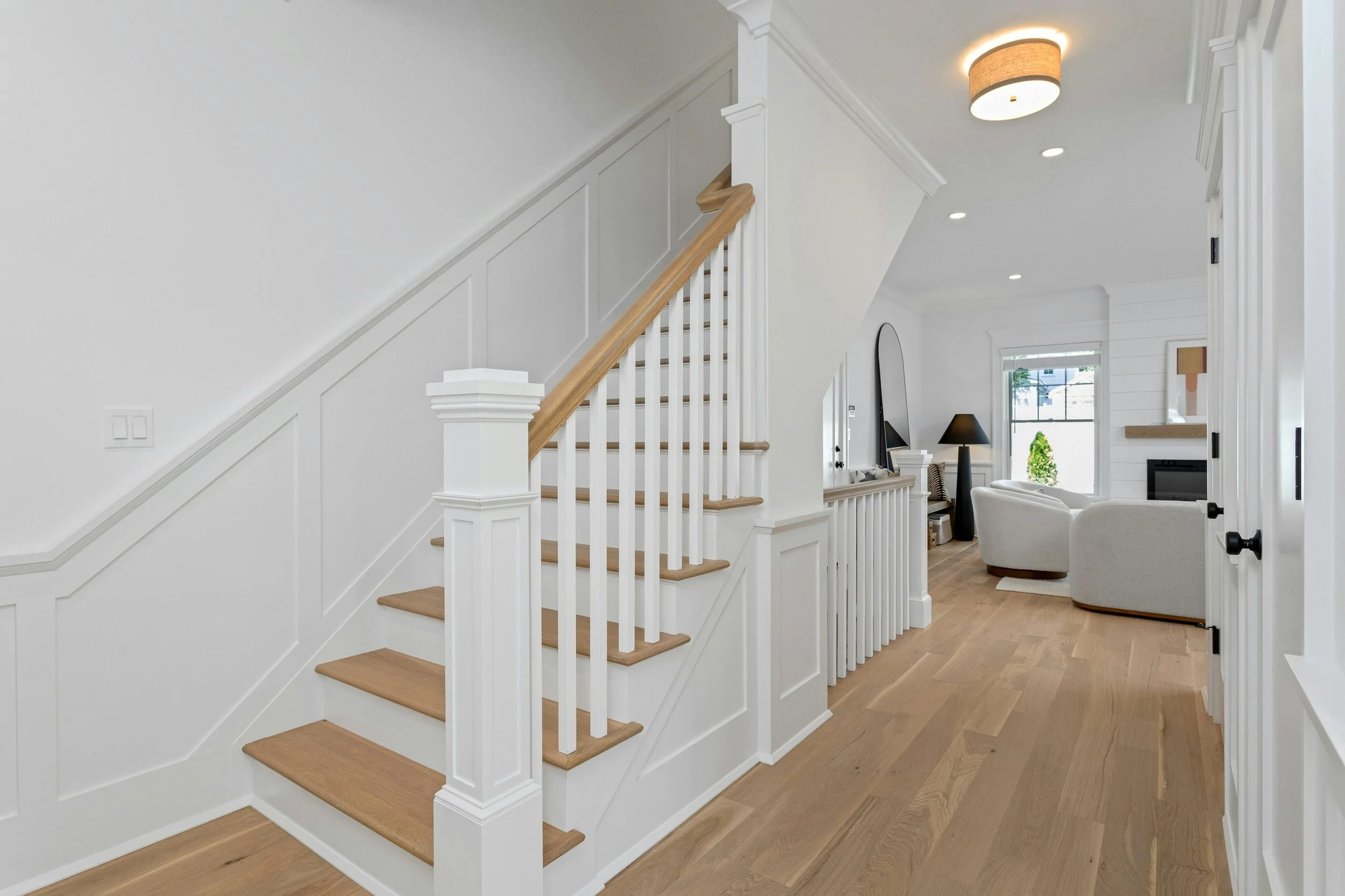 Interior view of a modern, white living room with a staircase featuring wooden steps and white spindles, leading upstairs. The room has light hardwood flooring, a couch, a black floor lamp, and a window with a small green plant outside. The ceiling h