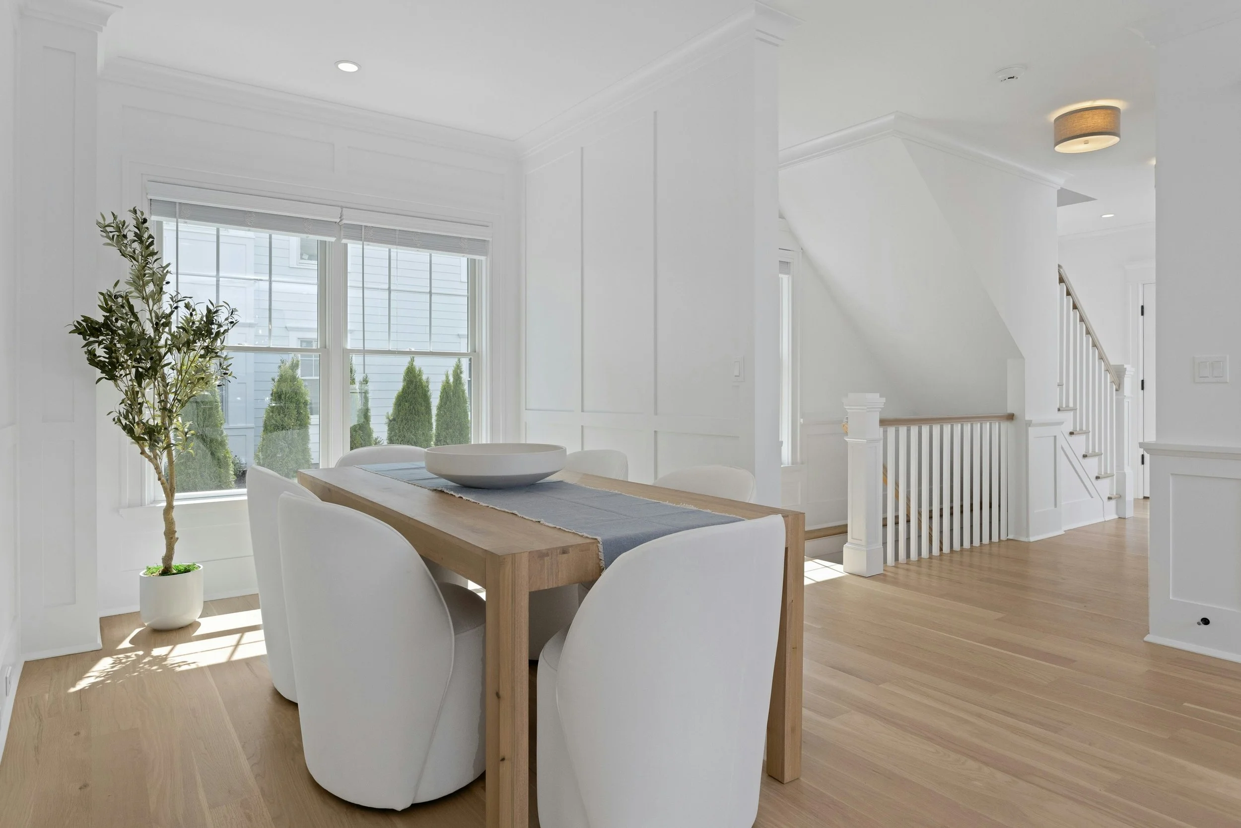 Bright dining area with white walls and light wooden floor, large window with blinds, indoor plant, wooden dining table with white chairs, decorative bowl, staircase with white railings, ceiling light fixture.