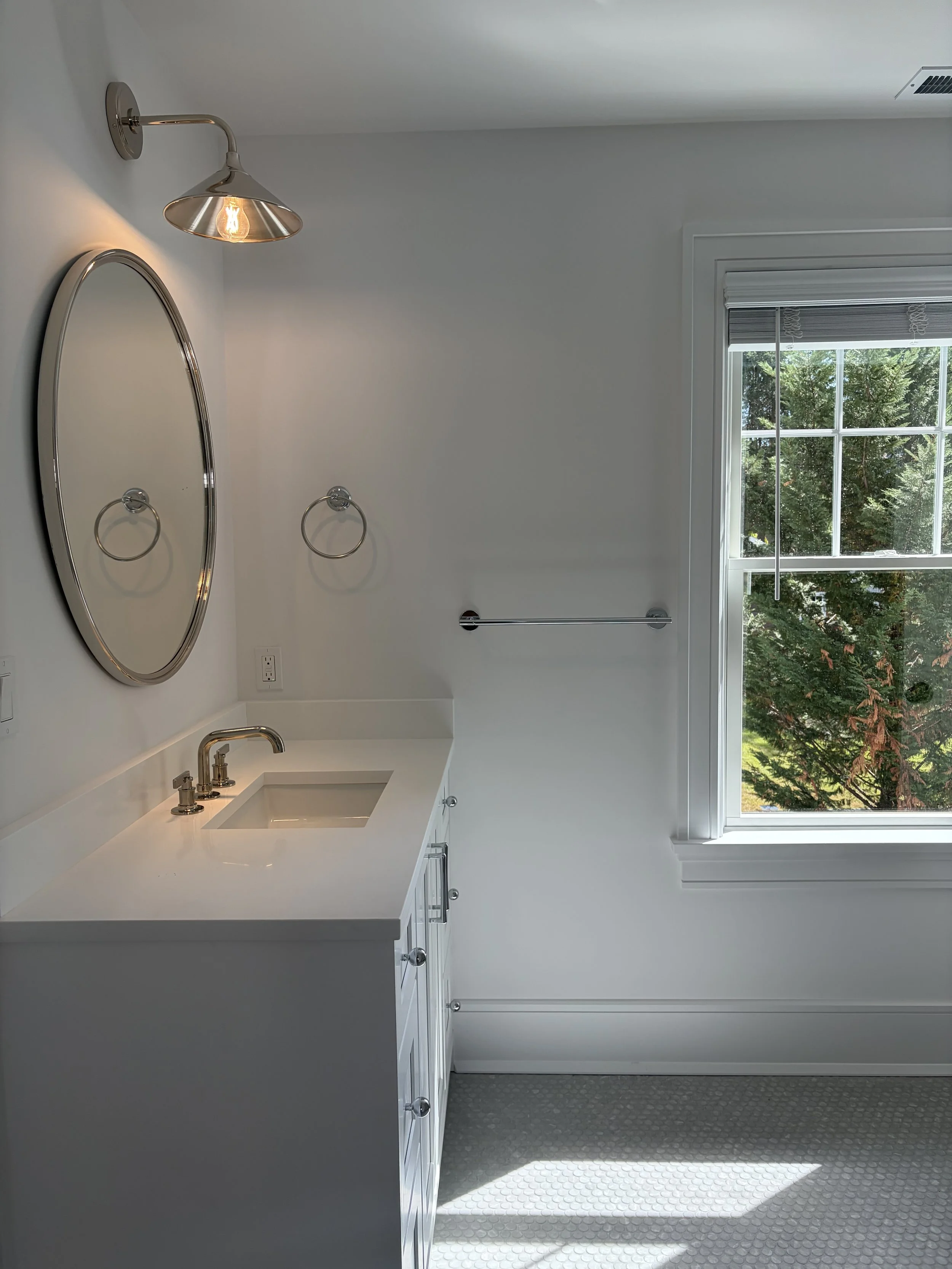 A bright, minimalist bathroom with a white vanity, a round mirror, a chrome wall light, and a window with a view of green trees outside.