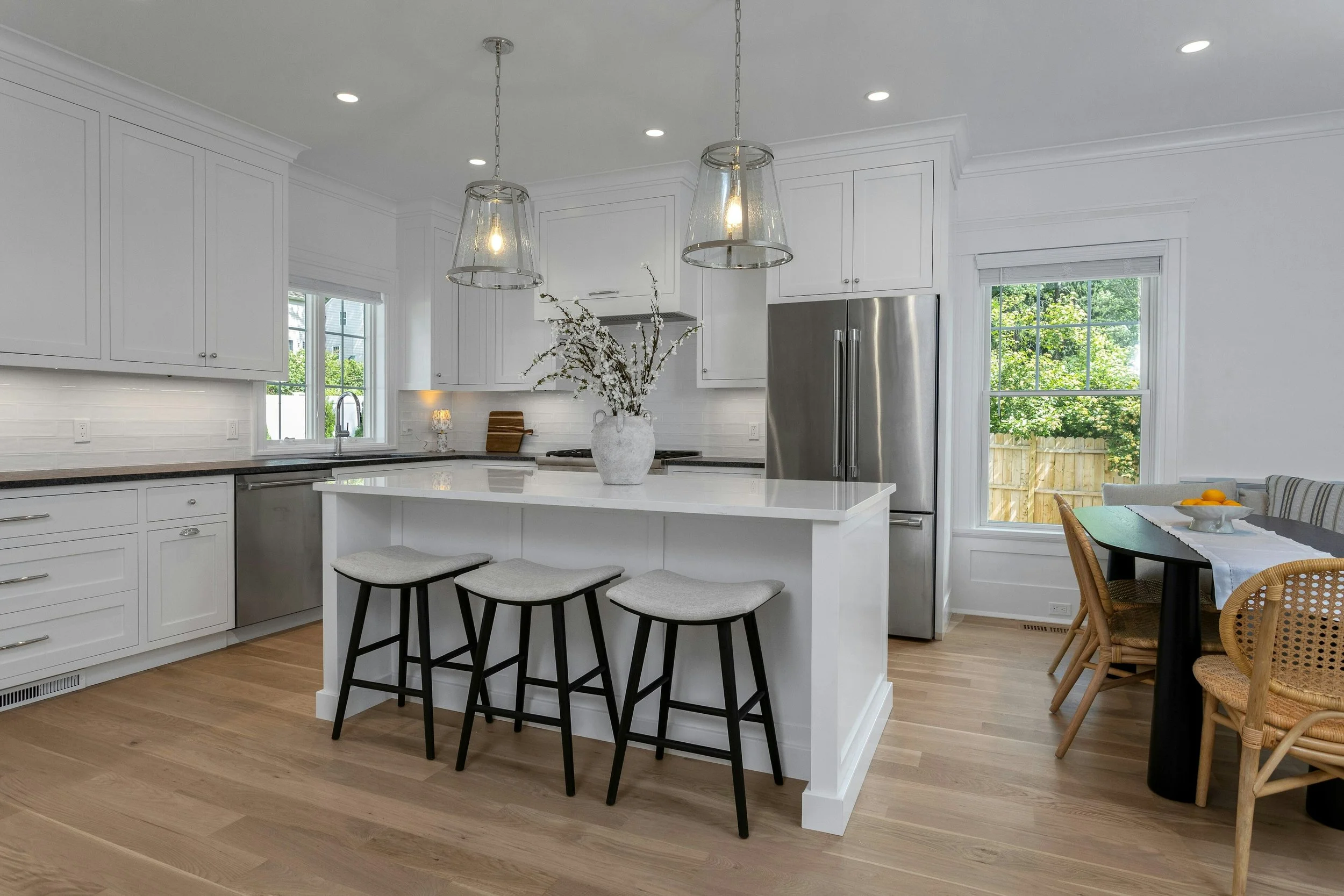 Modern white kitchen with a large island, black stools, and stainless steel appliances. Decor includes a white vase with branches, located on the island. There are large windows bringing in natural light with a view of a backyard with trees and a woo