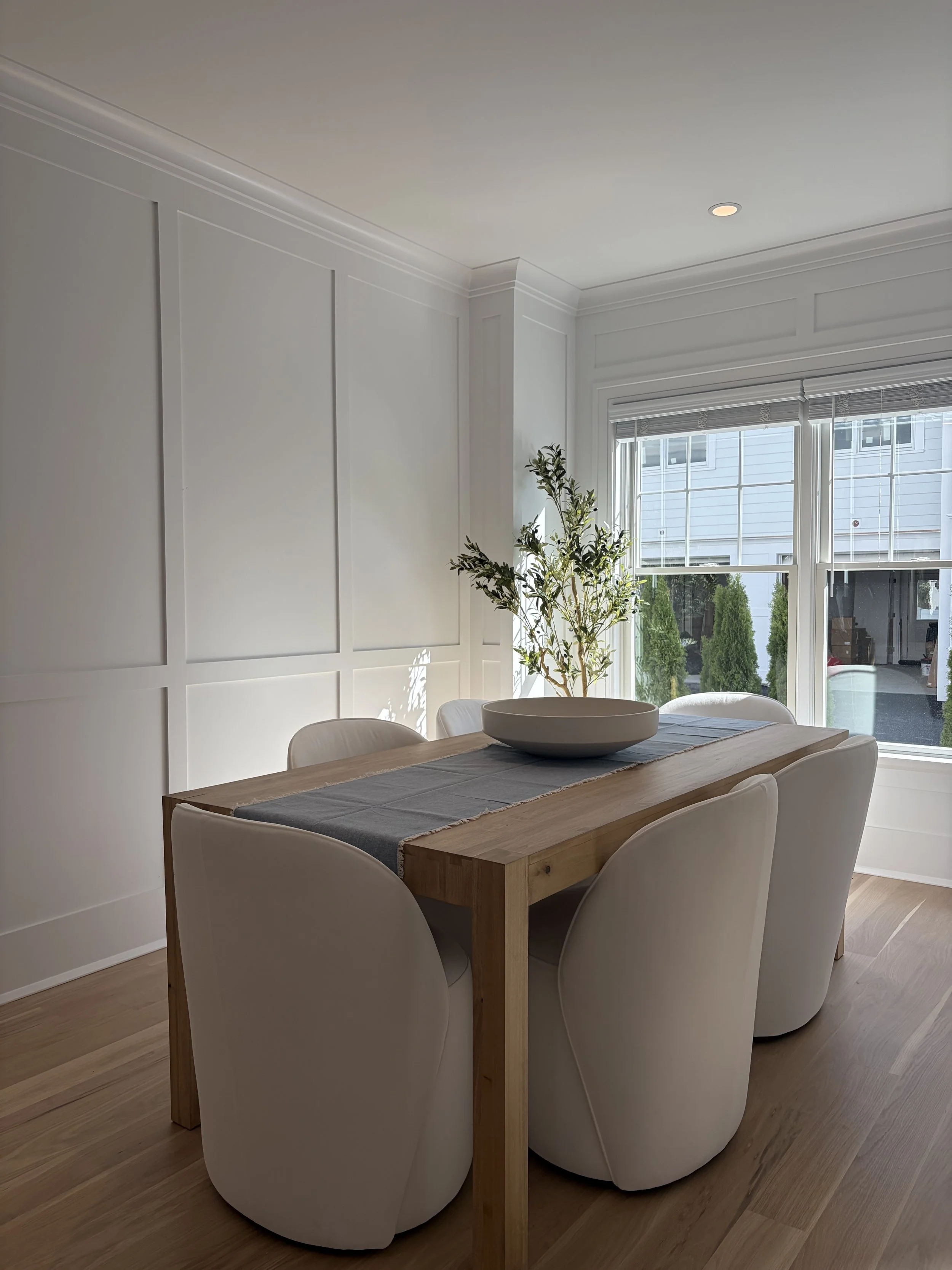 A dining room with a wooden table, six cream-colored chairs, a decorative bowl on a runner, a large window with blinds, and a potted plant in the corner, with white paneled walls and a ceiling with crown molding.