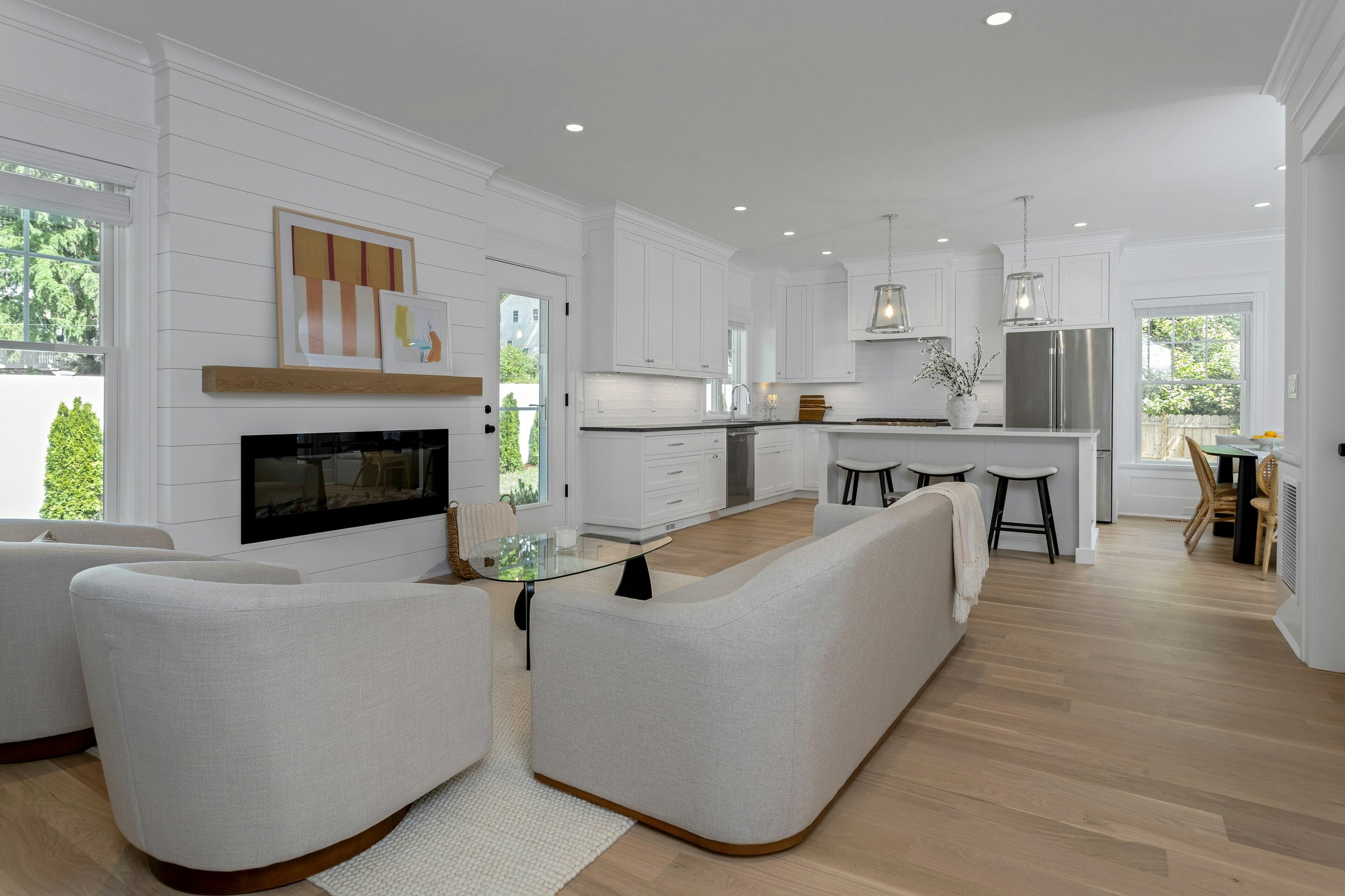 Bright, modern kitchen with white cabinets, a kitchen island with barstools, pendant lights, stainless steel appliances, and large windows bringing in natural light.