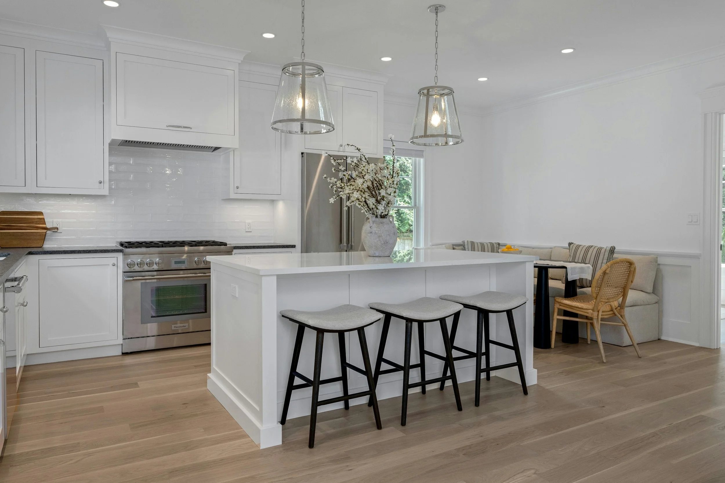 A modern kitchen with white cabinets, a stainless steel stove, a white kitchen island with a vase of white flowers, and four black bar stools. There's a dining area with a beige sofa and a table with some yellow fruit in the background. Two pendant l