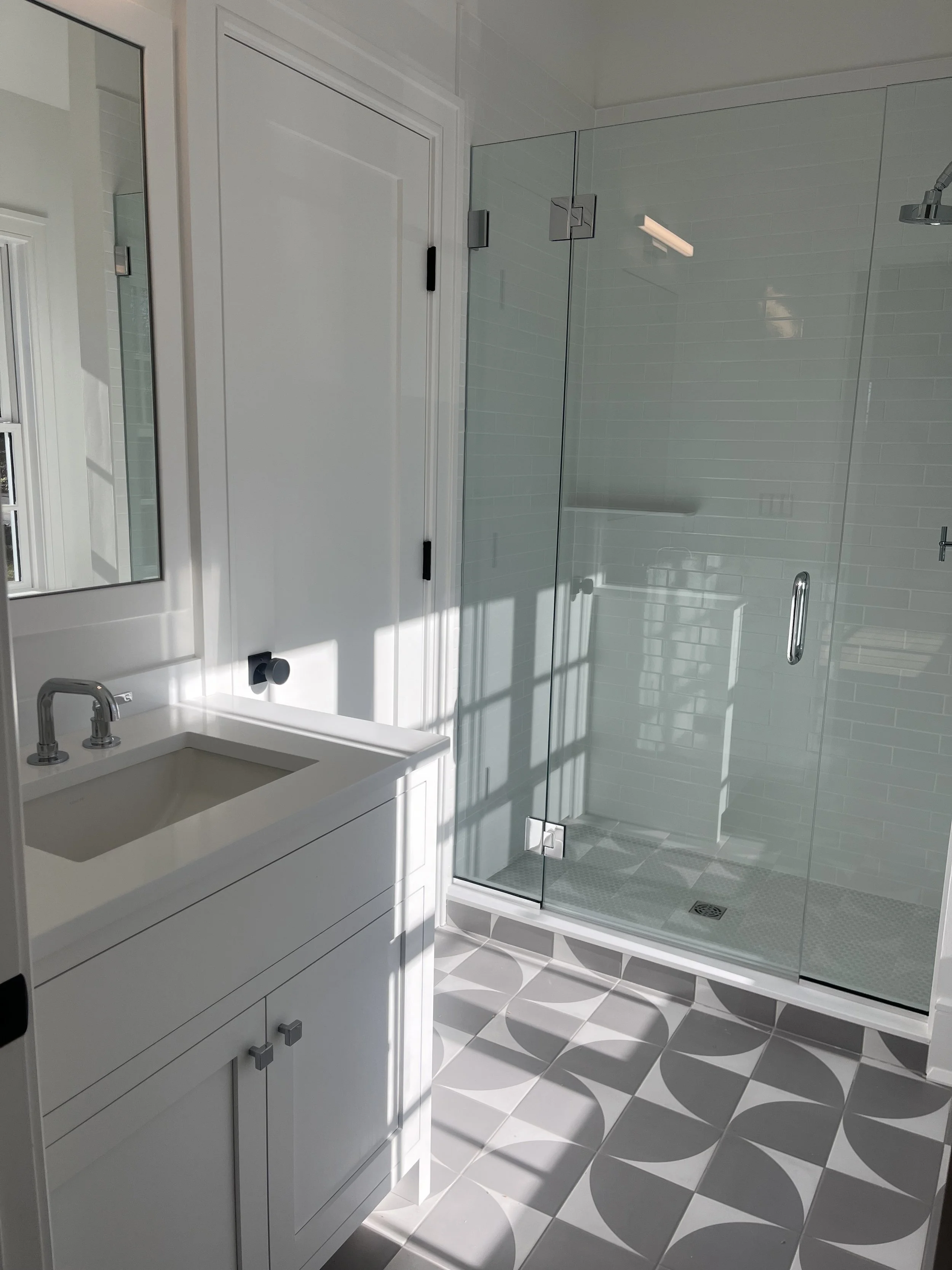 A modern bathroom featuring a white vanity with a sink, a mirror, and a clear glass walk-in shower with a chrome handle, white tiled wall, and geometric patterned floor tiles.