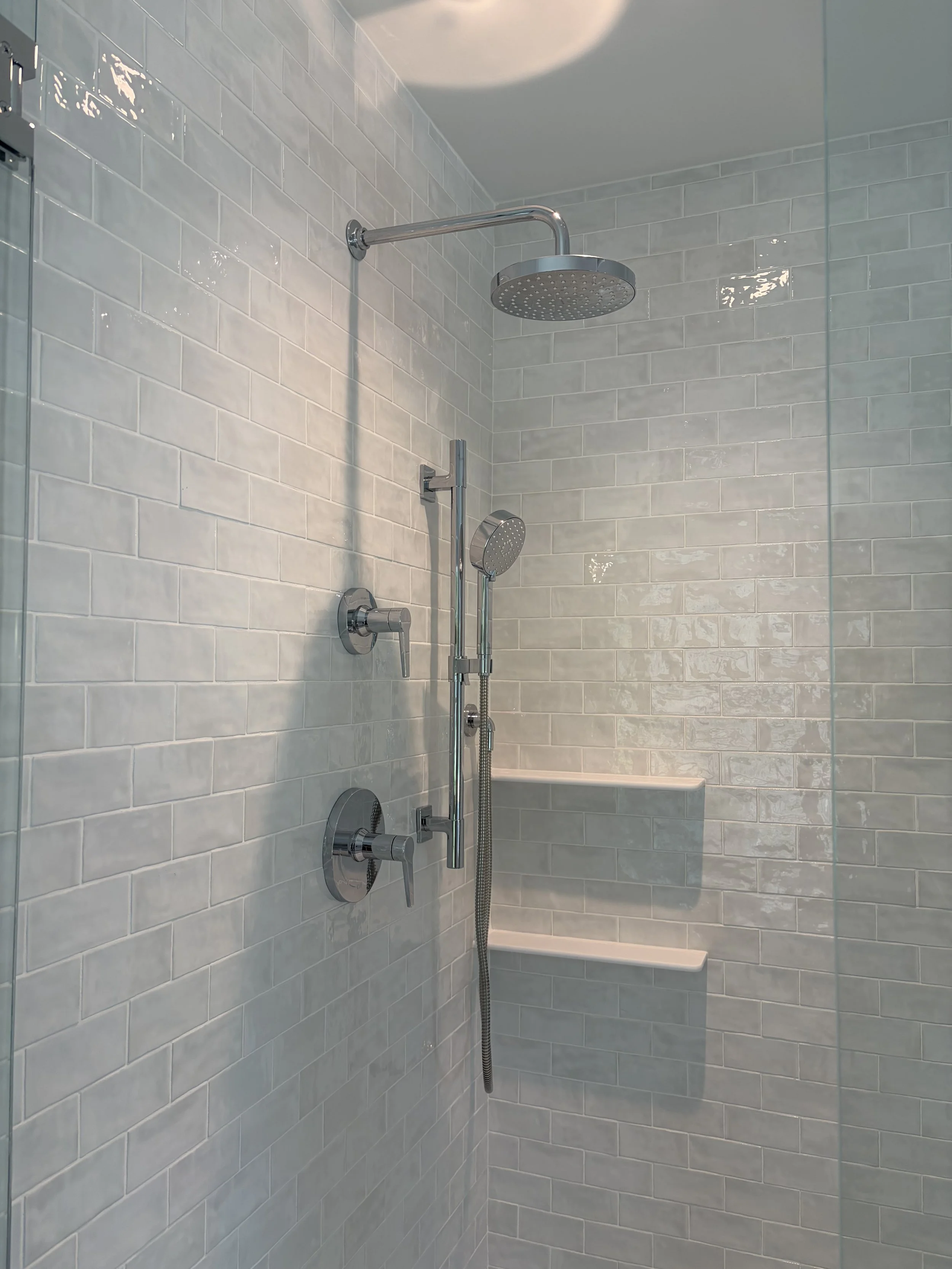 A modern shower area with two showerheads, one large overhead rain shower and a handheld shower, mounted on a white tiled wall.