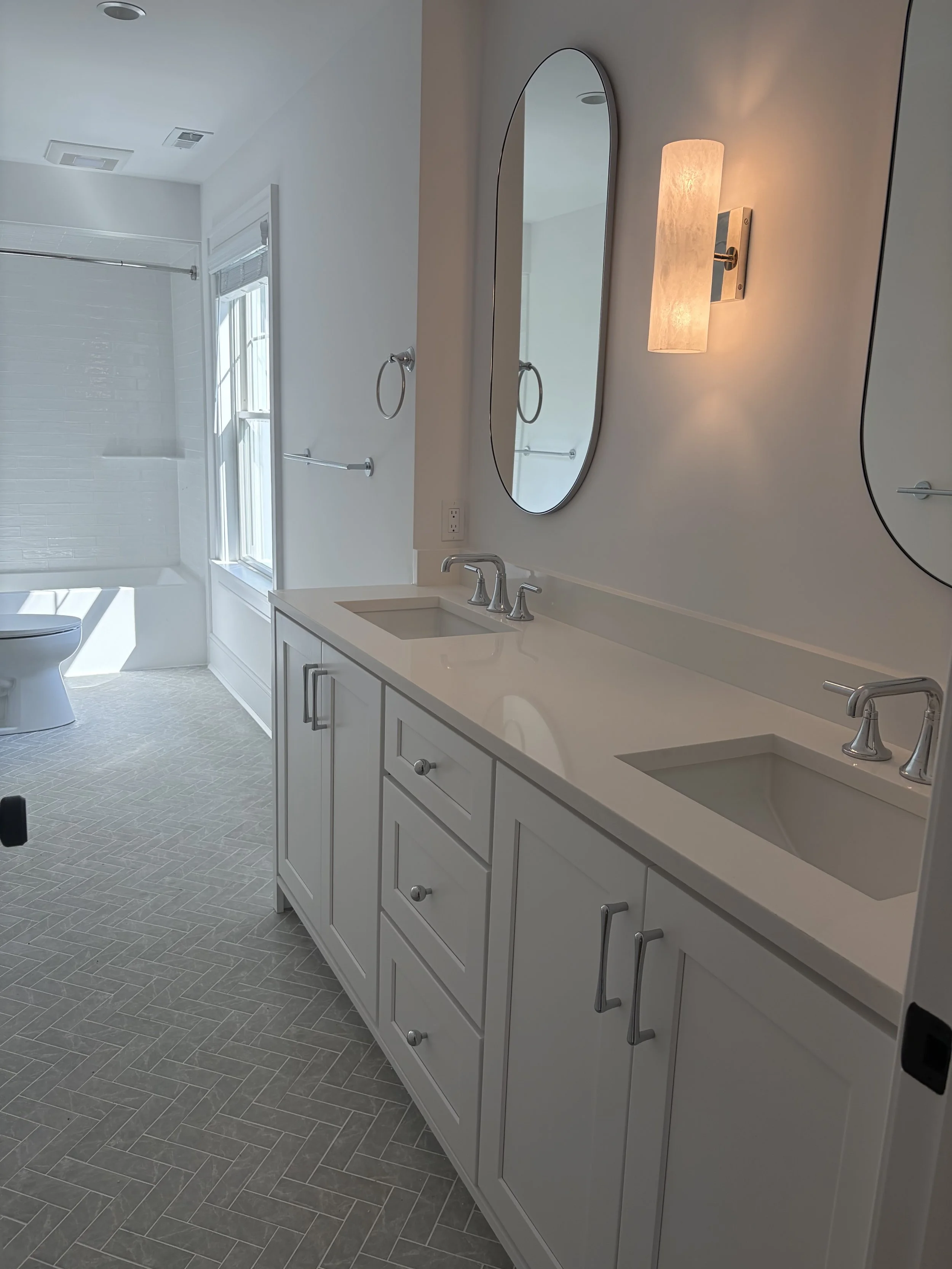 A modern bathroom with white cabinetry, a double sink, large oval mirror, wall-mounted light, and a gray tile floor. A shower and toilet are visible in the background.