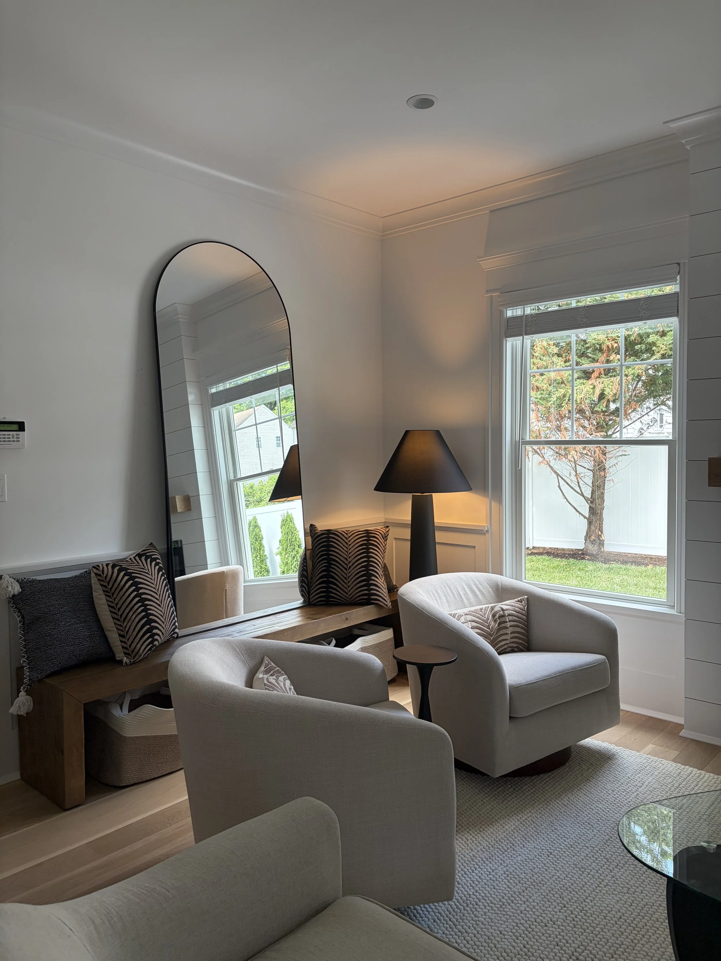 A cozy living room with a large window showing a tree outside, a white armchair, a black table lamp, a wooden bench with decorative pillows, a large mirror leaning against the wall, a patterned chair, and a glass coffee table on a textured rug.