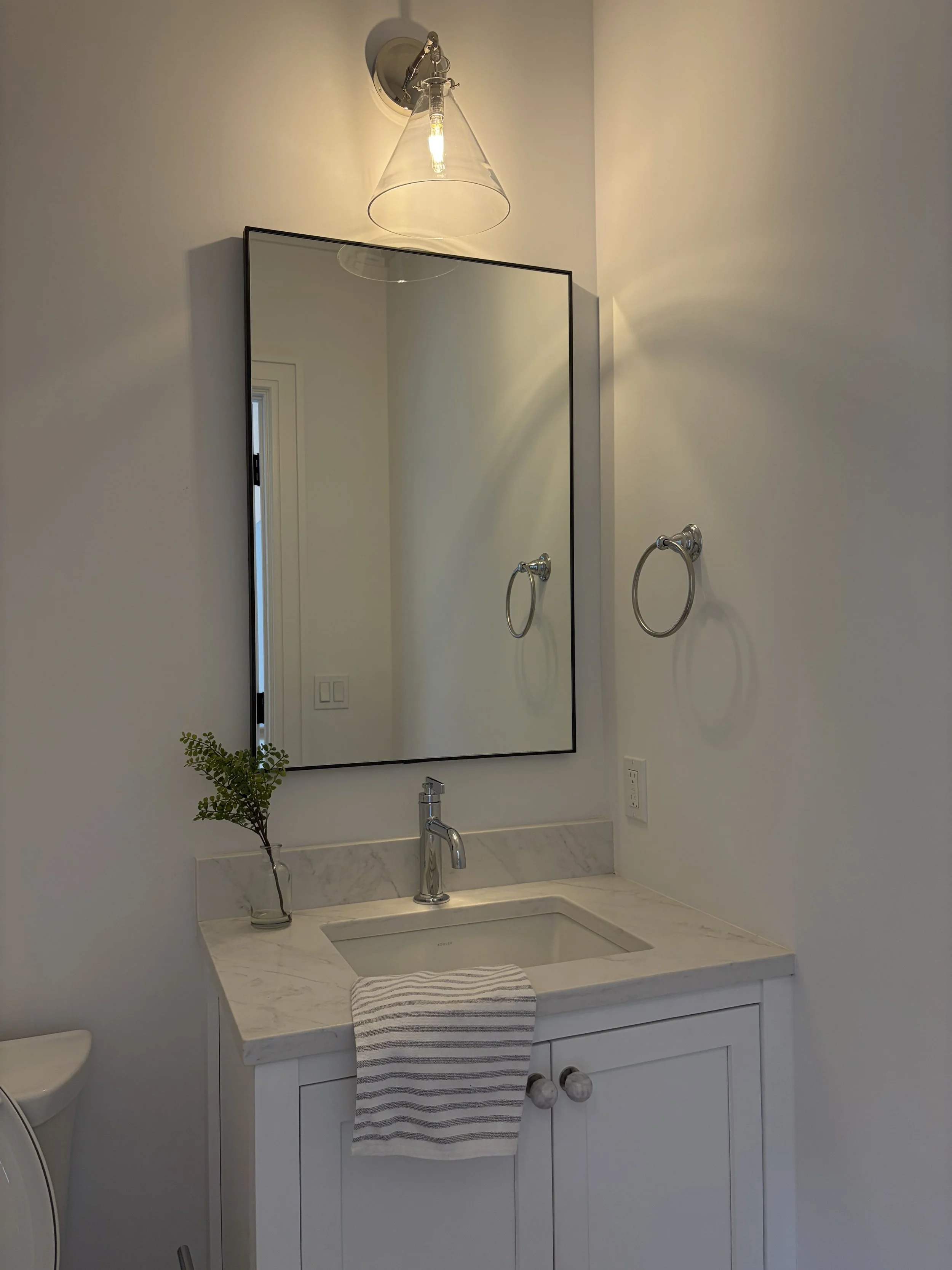 Bathroom vanity with marble countertop, white cabinet, rectangular mirror, wall-mounted towel ring, small plant in a glass vase, striped hand towel, wall light fixture with clear glass shade, and a window reflected in the mirror.