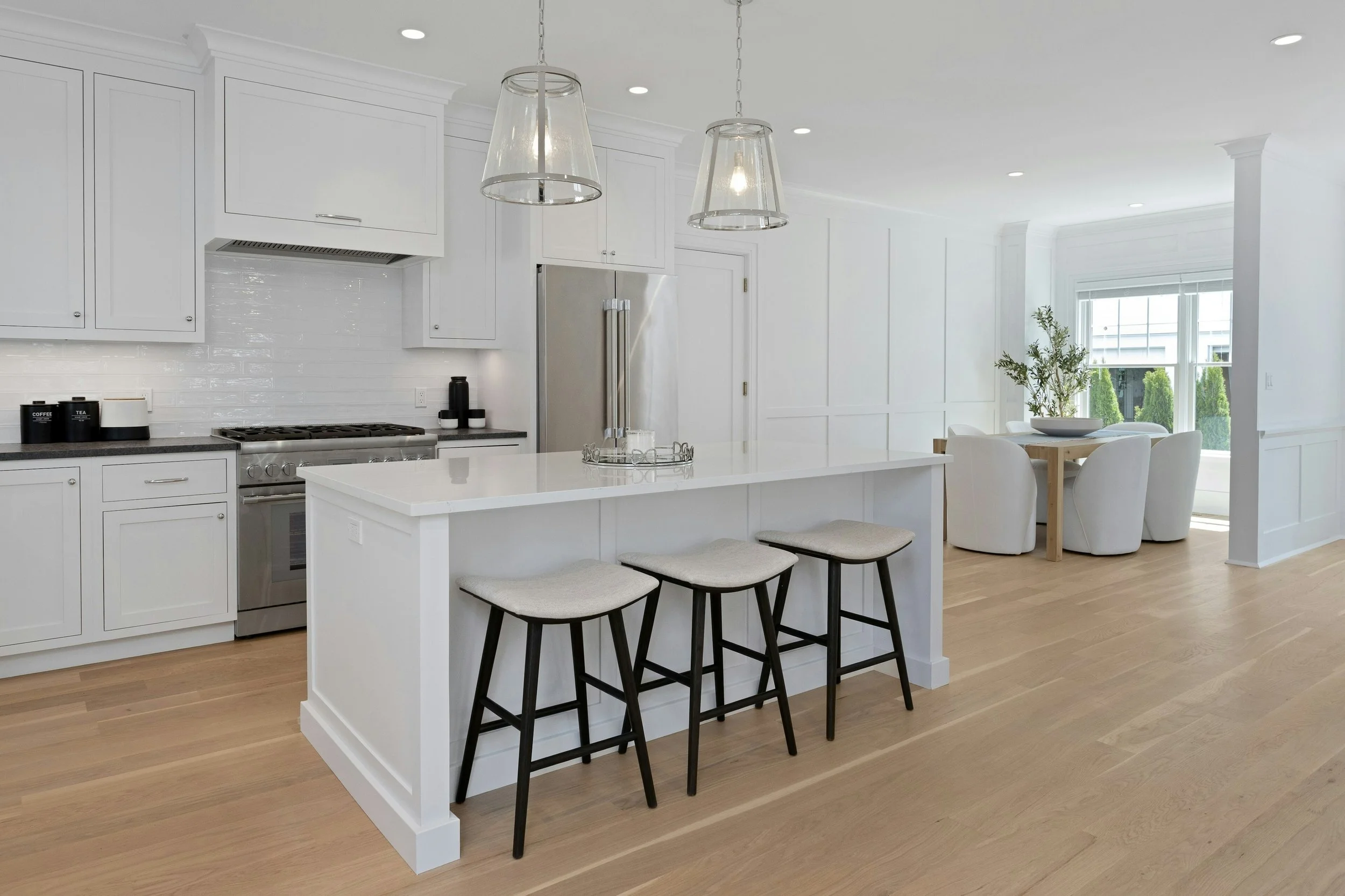 Modern white kitchen with an island, black stools, stainless steel appliances, and a dining area with a wooden table and white chairs near large windows.