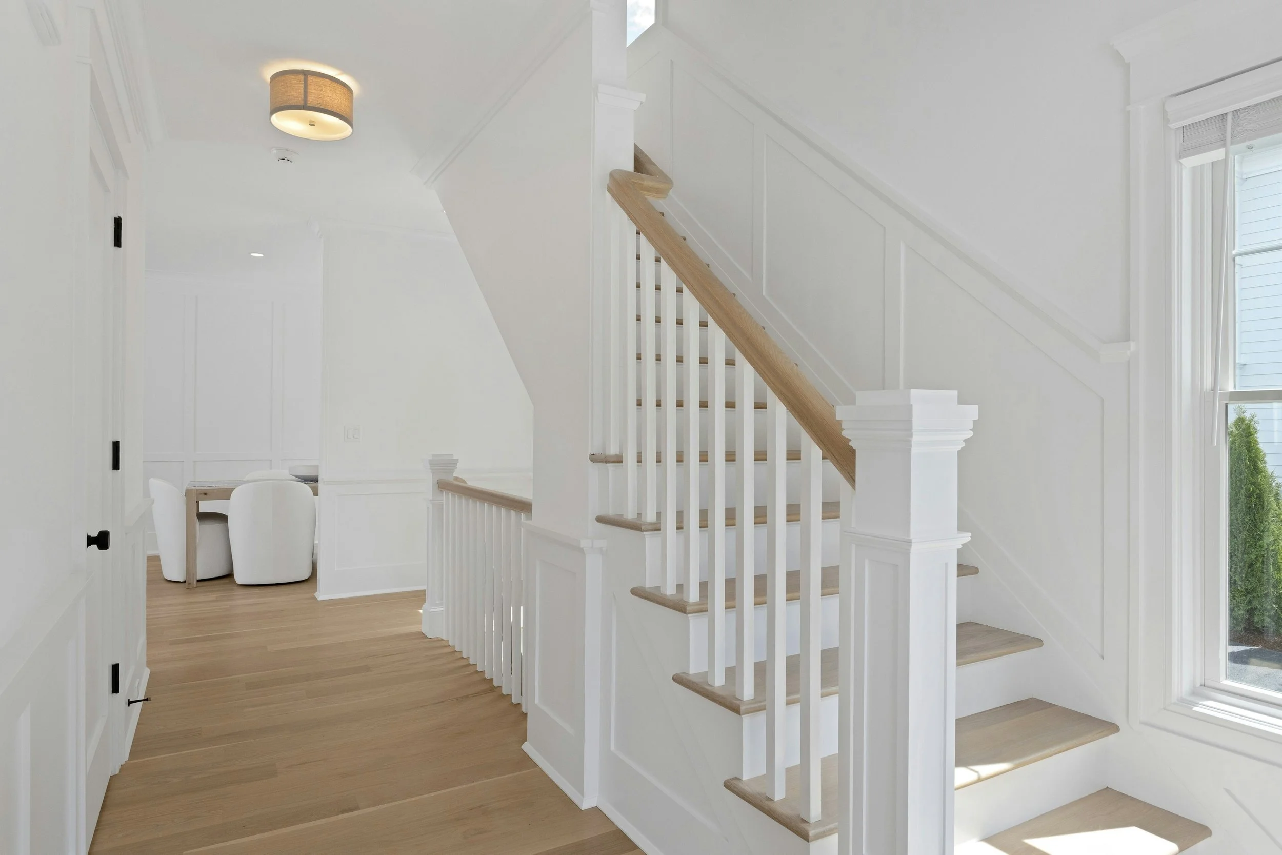 Interior view of a bright, white staircase with wooden handrails and balusters, with large windows allowing natural light in, and a partial view of a dining area with white chairs and a wooden table.