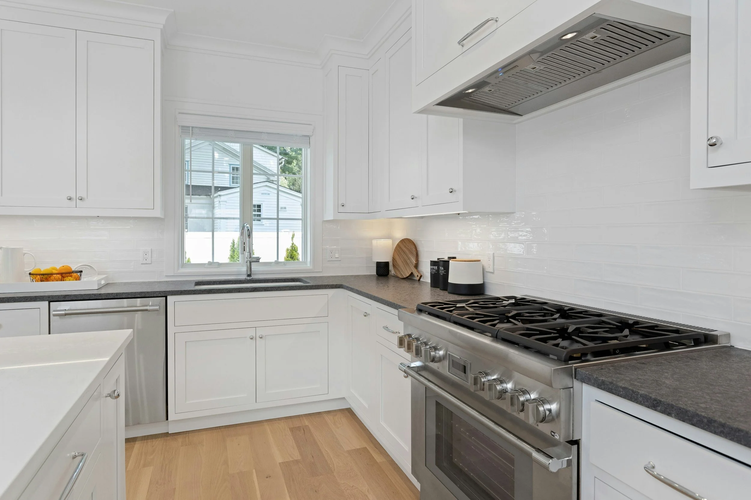 A modern kitchen with white cabinets, stainless steel appliances including a stove and exhaust hood, a window overlooking a residential neighborhood, and a black countertop with decorative items and a bowl of fruit.