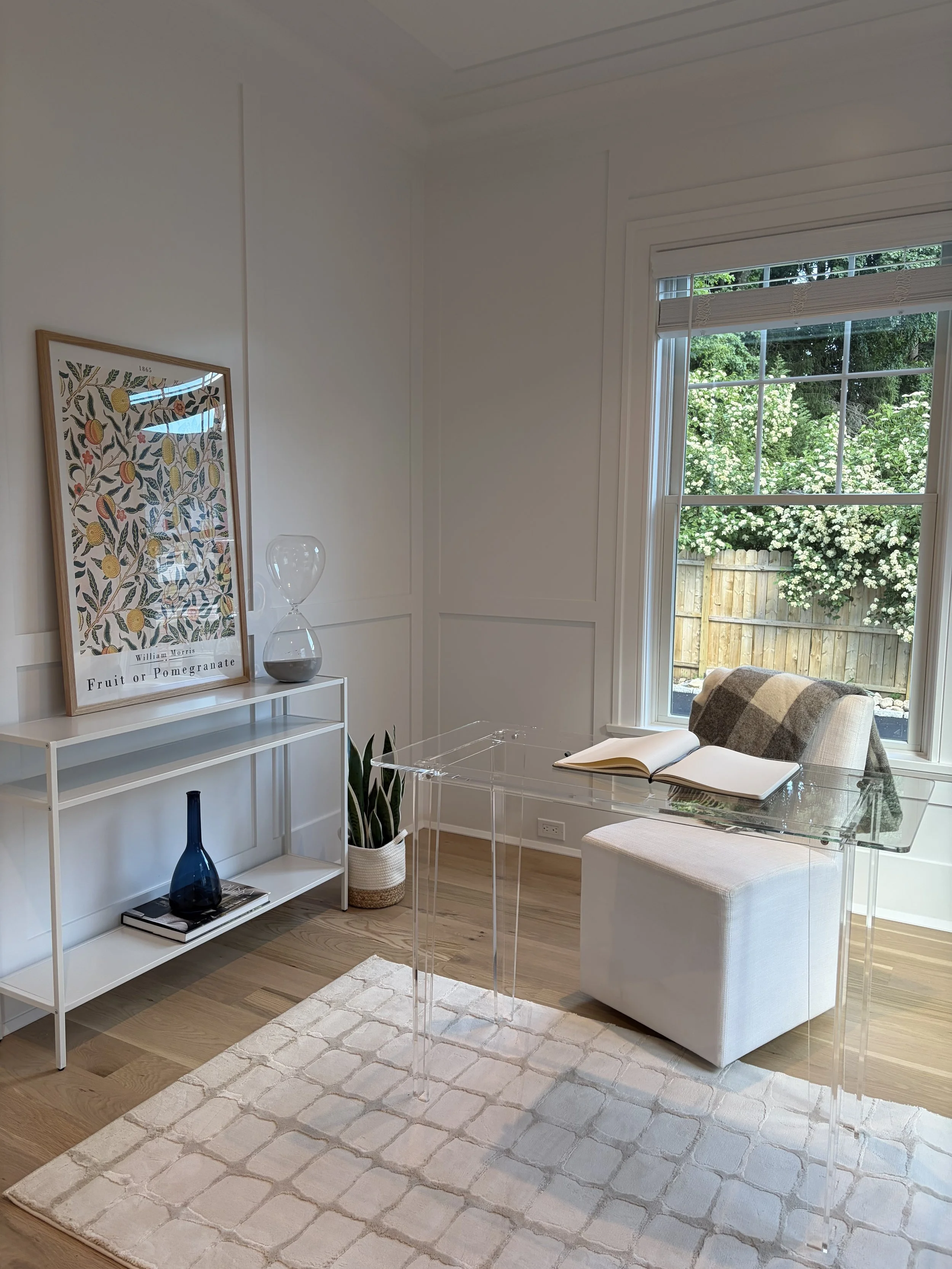 A cozy corner in a bright room with a large window showing greenery outside. There is a white shelf with decorative items, a glass hourglass, a potted plant, a window with a white blind, a plaid armchair, a clear acrylic side table with an open book,