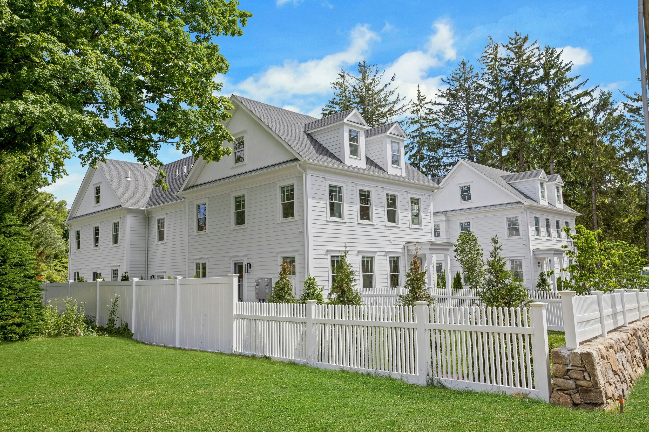 A white multi-story house with a gray roof, surrounded by a white picket fence and green lawn. Tall trees and a blue sky with some clouds are in the background.